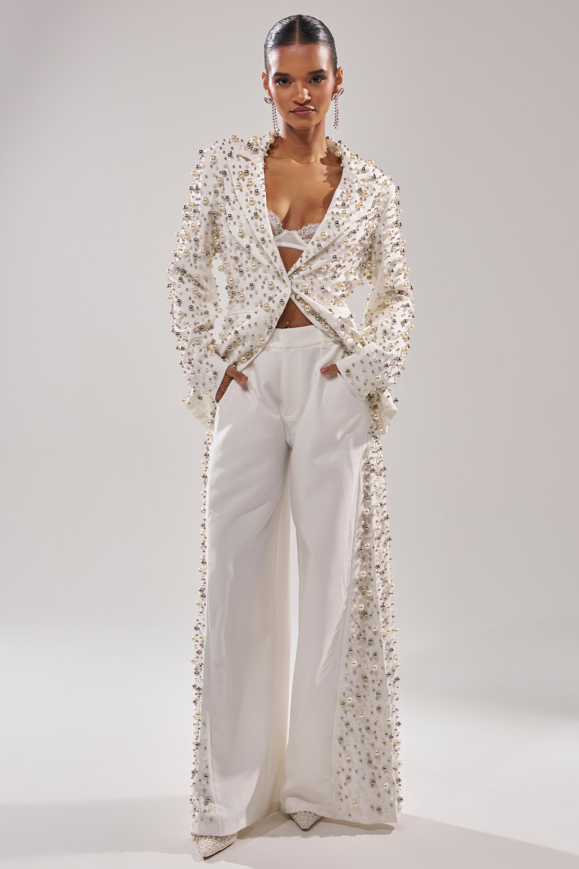 A woman models the KEEP IT GOING EMBELLISHED BLAZER in white, paired with wide-leg trousers and a matching bralette. Her hair is slicked back as she poses with hands in her pockets against a plain background.