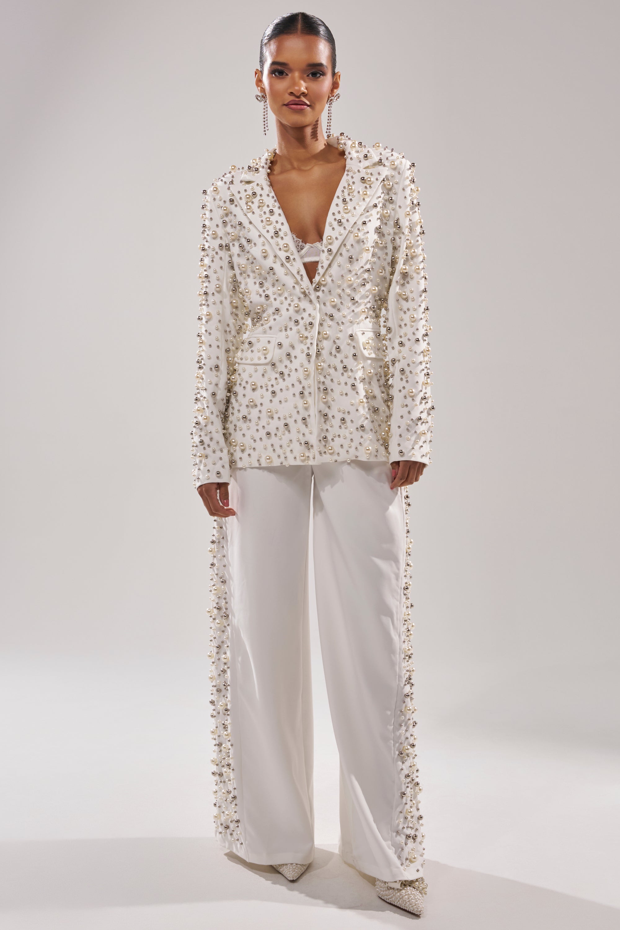 A model wears the KEEP IT GOING EMBELLISHED BLAZER with matching wide-leg pants and white pointed heels, featuring intricate pearl and bead detailing against a plain background.