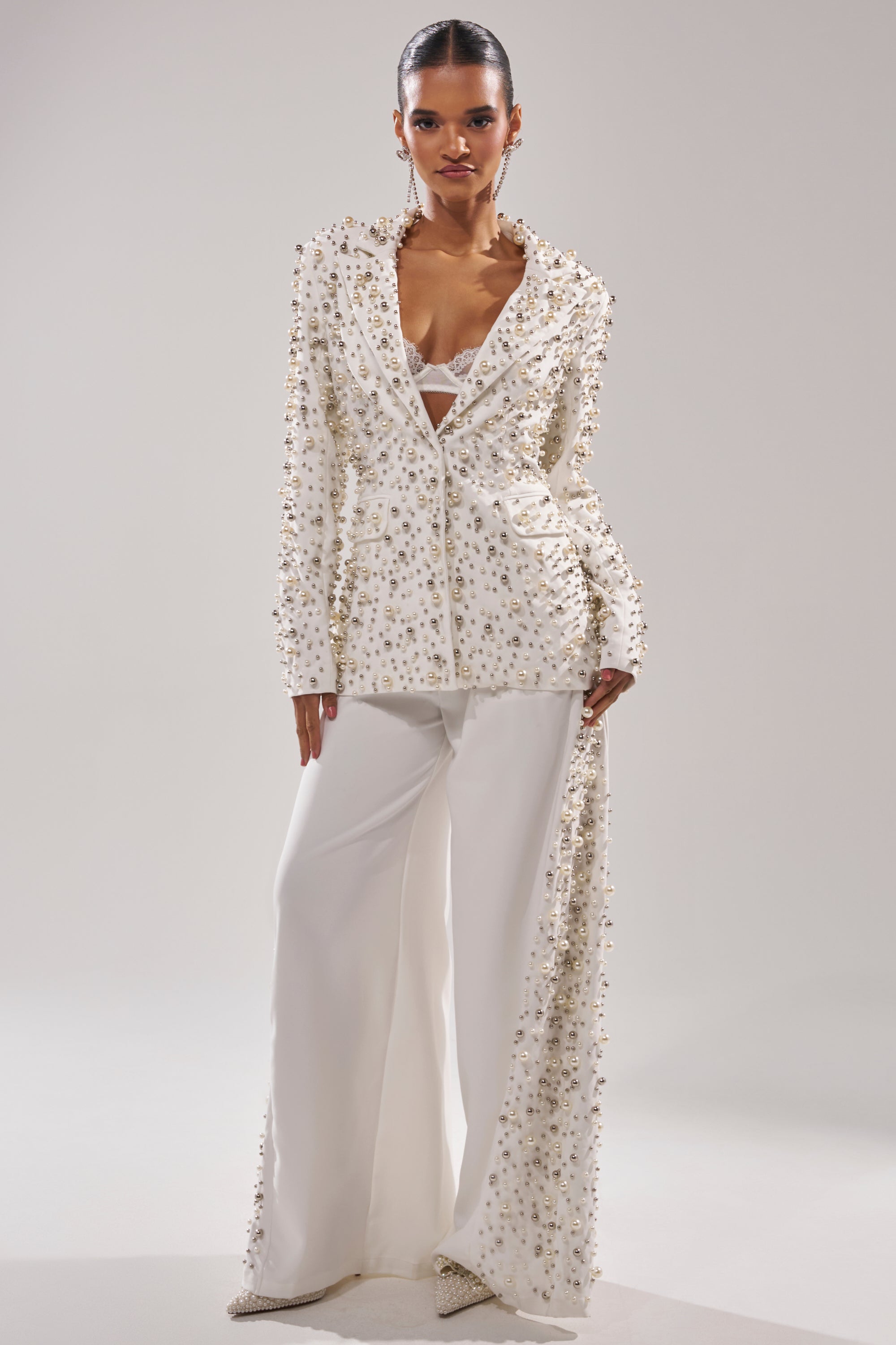 A woman stands confidently in the KEEP IT GOING EMBELLISHED BLAZER, a stylish white jacket with sparkling details, wide-leg pants, and a dramatic train, paired with a white bralette against a plain light background.