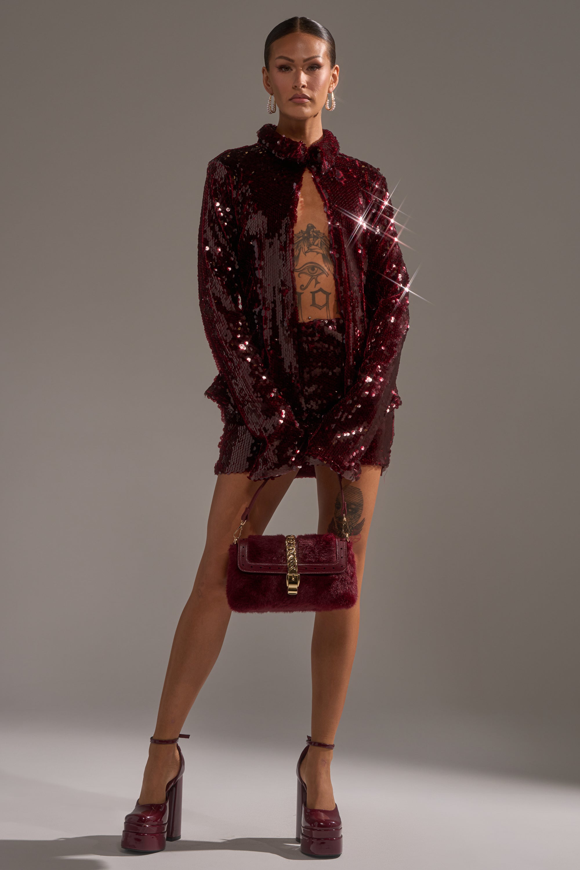 A woman poses confidently in the SHINING DOWN ON YOU SEQUIN MINI SKIRT and a matching burgundy blazer, revealing tattoos on her legs and torso. She accessorizes with a burgundy handbag and high-platform heels against a plain gray background.