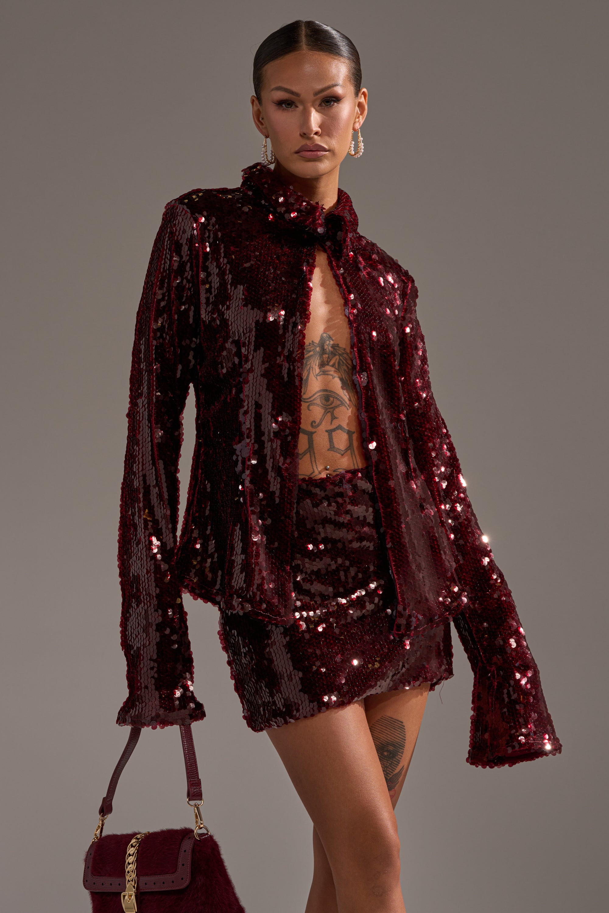 A person poses confidently in a shiny, dark red SHINING DOWN ON YOU SEQUIN BUTTON DOWN jacket and mini skirt, with a matching handbag, hoop earrings, and visible tattoos—an ideal look for holiday parties.