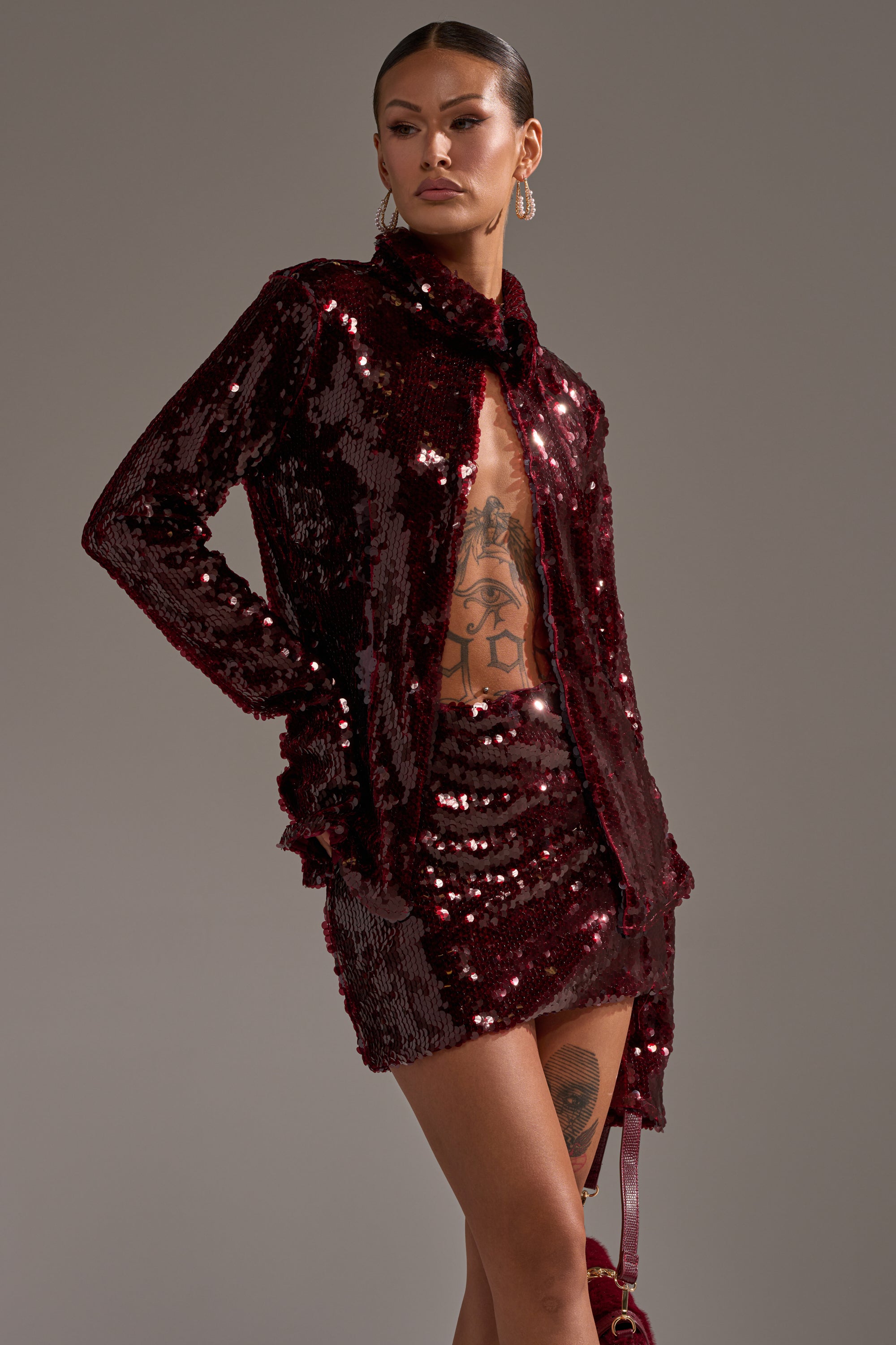 Wearing the SHINING DOWN ON YOU SEQUIN BUTTON DOWN in dark red with a matching mini skirt over a mesh top, this person shows off their tattooed torso, accessorized with slicked-back hair, hoop earrings, and a small matching item against a neutral backdrop.