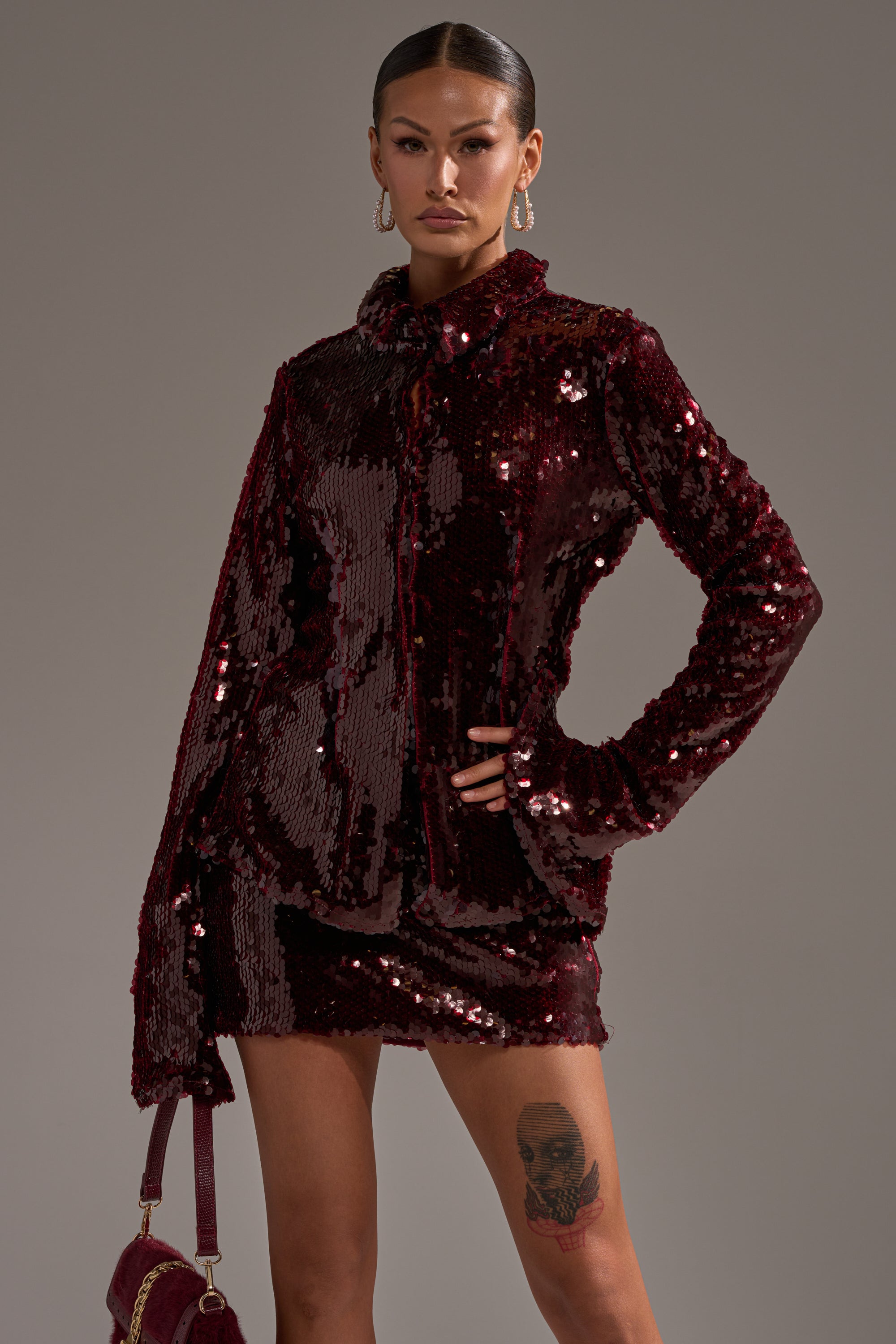 A woman stands confidently in the SHINING DOWN ON YOU SEQUIN BUTTON DOWN jacket and matching mini skirt, holding a small bag. She has sleek hair, hoop earrings, a face tattoo on her thigh, and poses against a plain gray background.