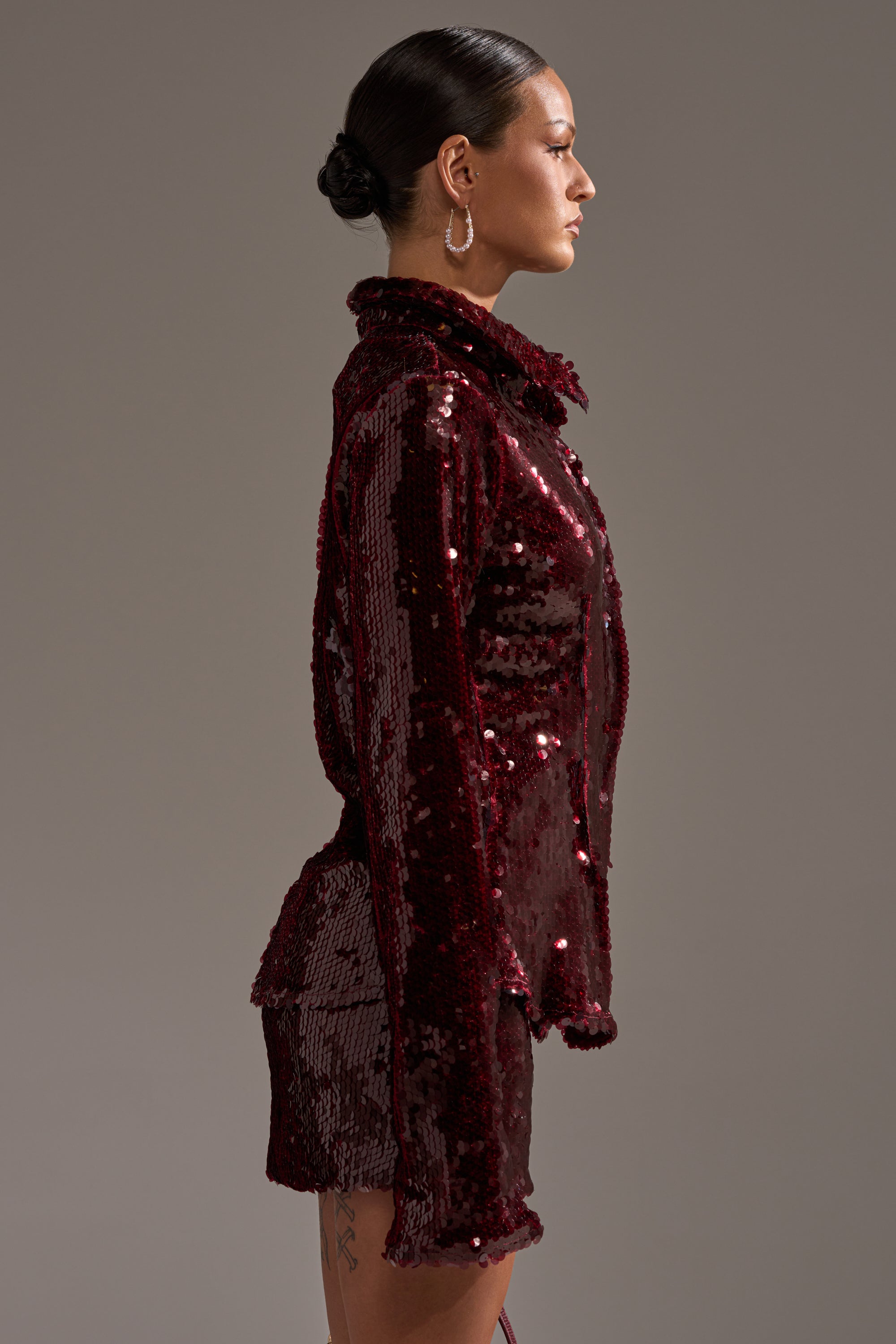 A woman in profile stands against a plain background, wearing the SHINING DOWN ON YOU SEQUIN BUTTON DOWN with matching shorts. Her hair is sleek and pulled into a low bun, complemented by large hoop earrings for a festive party look.