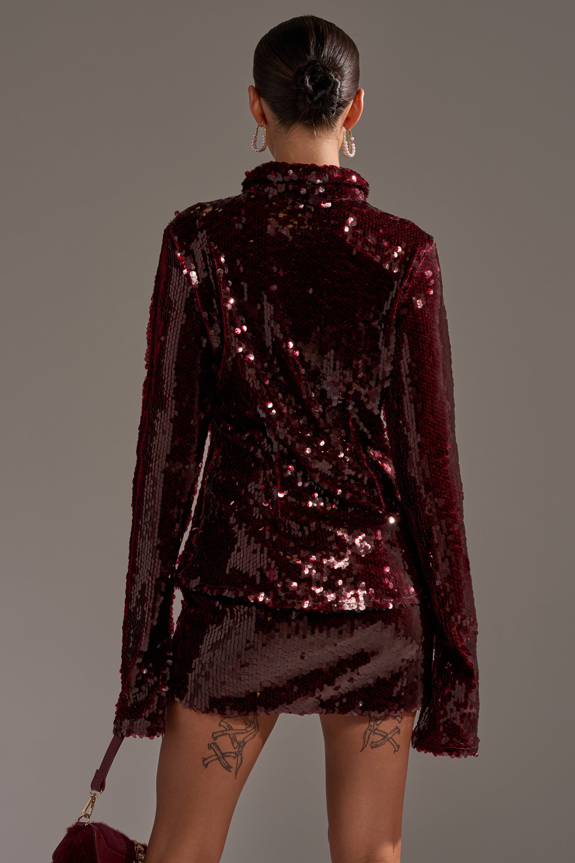 A woman with her hair in a bun is seen from behind, wearing the SHINING DOWN ON YOU SEQUIN BUTTON DOWN and matching mini skirt, holding a small handbag. She has tattoos on the backs of her thighs.