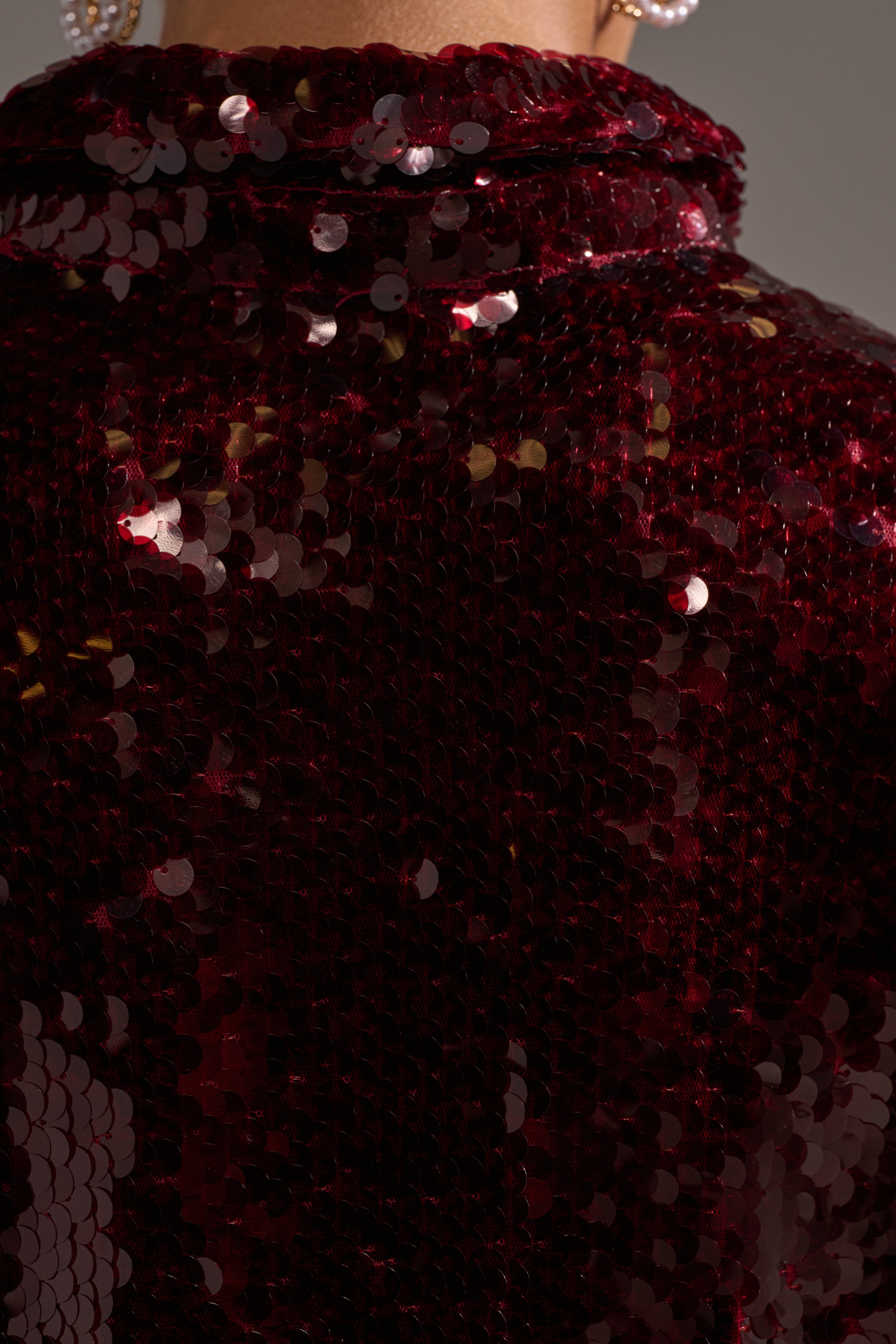 Close-up of the SHINING DOWN ON YOU SEQUIN BUTTON DOWN, covered in shiny, dark red sequins that catch the light. The textured, shimmering surface stands out, with a hint of a person's neck and pearl earring at the top edge.