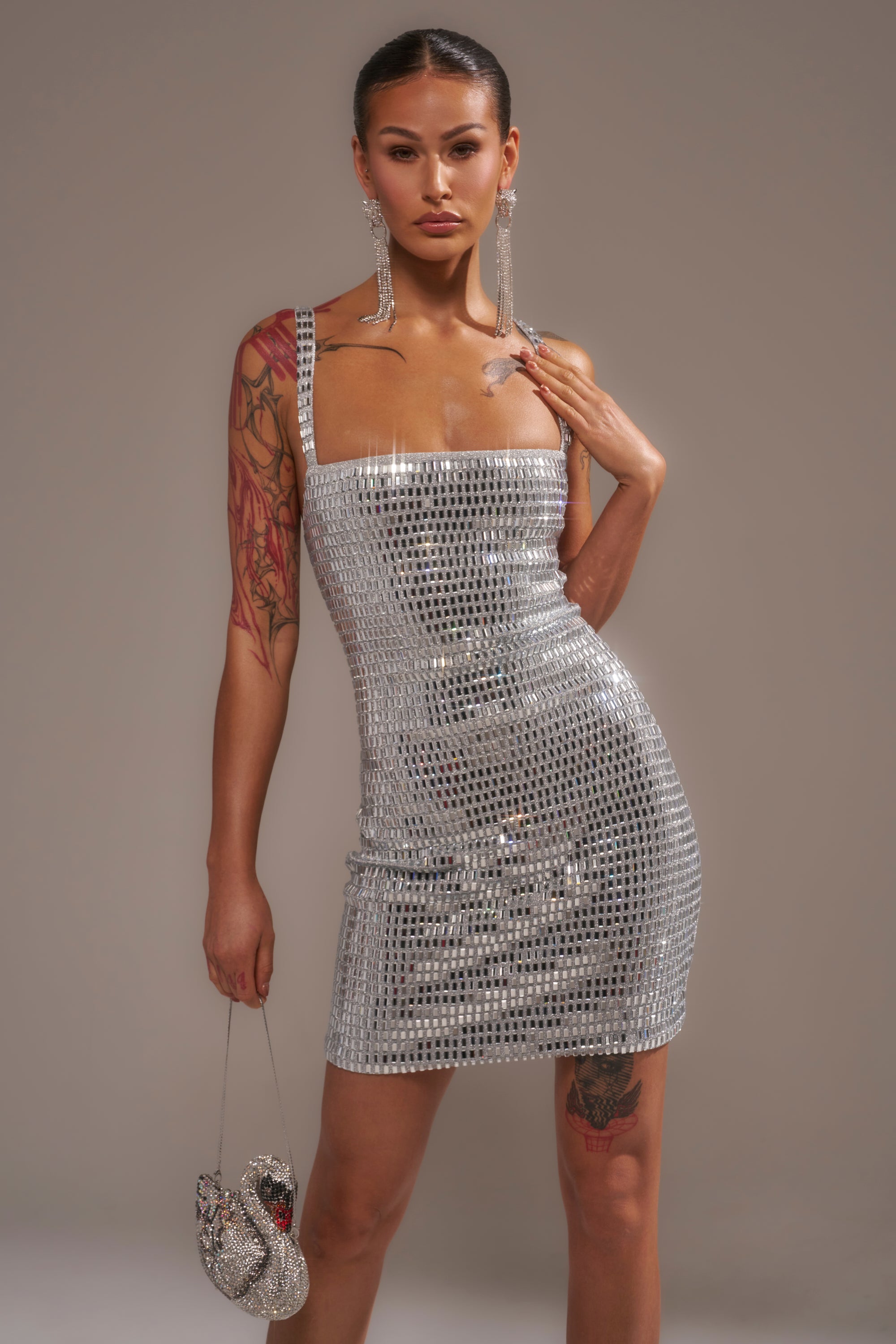 A woman poses in the BAILEY RHINESTONE MINI DRESS, a sleeveless silver sequin party dress, holding a swan-shaped handbag. She accessorizes with long earrings and shows visible tattoos, standing against a plain neutral background.