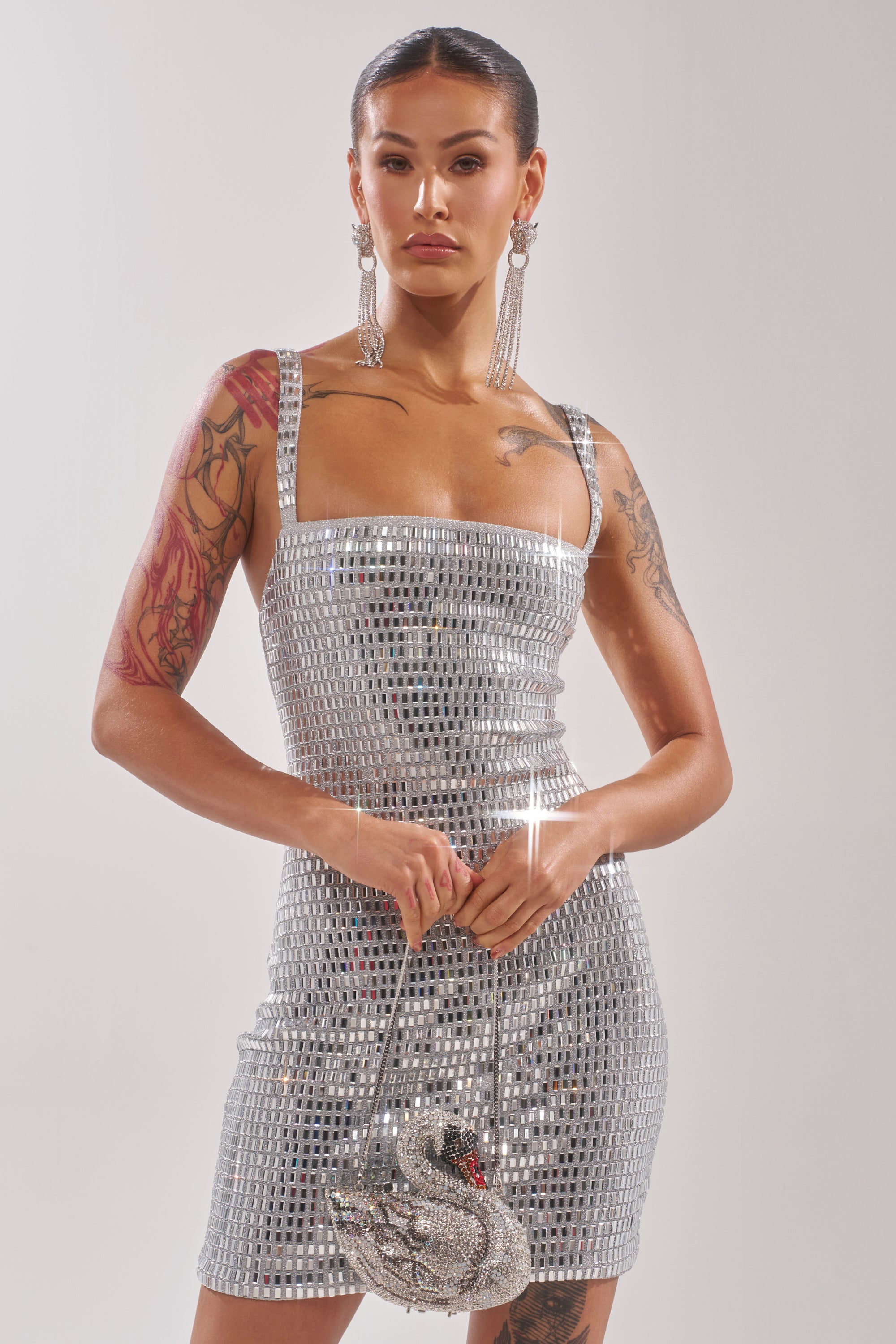 A woman stands confidently in the BAILEY RHINESTONE MINI DRESS, its mirror sequins catching the light. She holds a swan-shaped handbag, accessorized with long earrings, visible tattoos, and sleek hair against a plain backdrop. 2