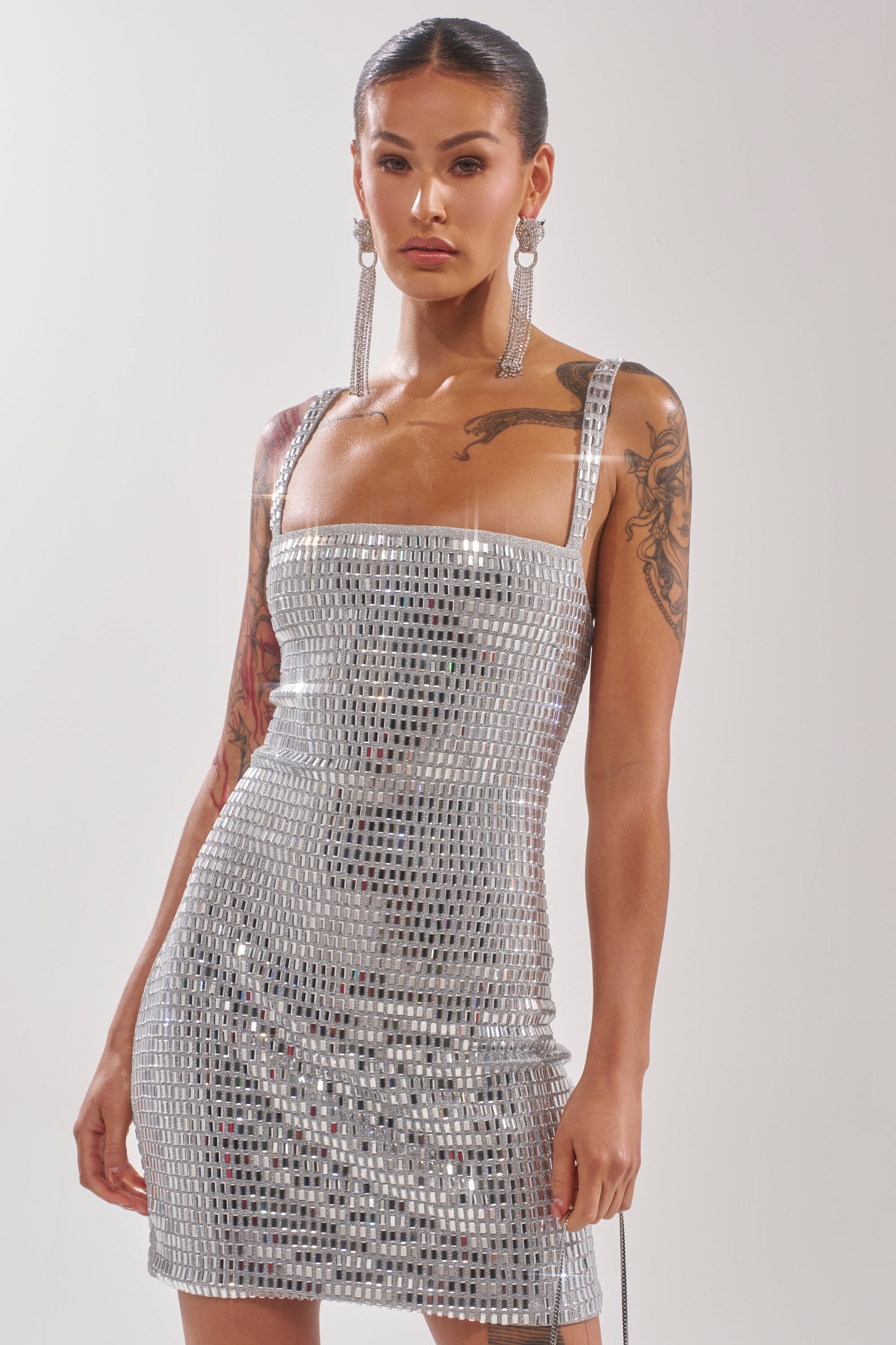 Wearing the BAILEY RHINESTONE MINI DRESS, a woman with tattoos on both arms and dark hair pulled back poses against a plain light background, pairing the sleeveless, square-neck silver sequin dress with long dangling earrings. 3