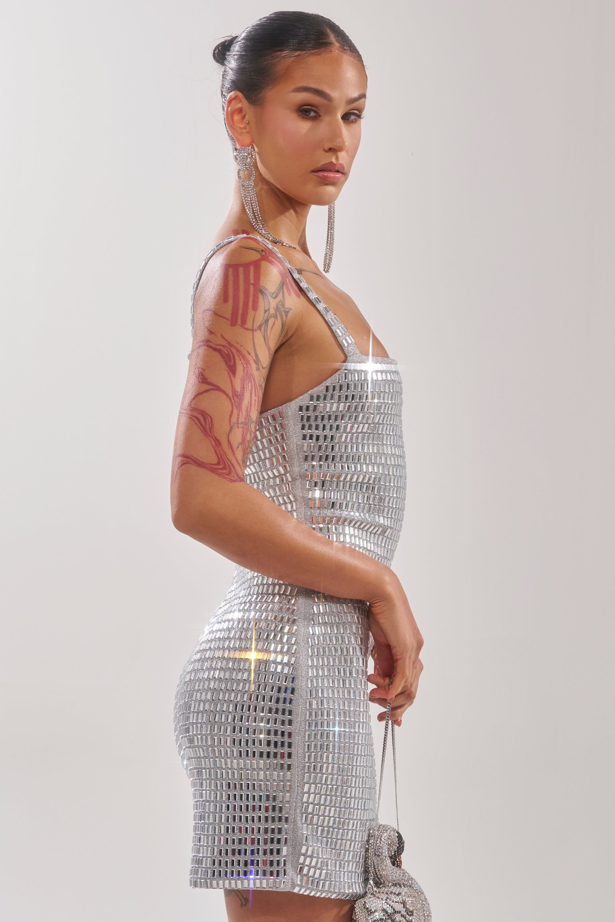 A woman with long earrings and visible arm tattoos poses in the BAILEY RHINESTONE MINI DRESS, a silver sequin mini dress, holding a matching handbag against a light background. 4