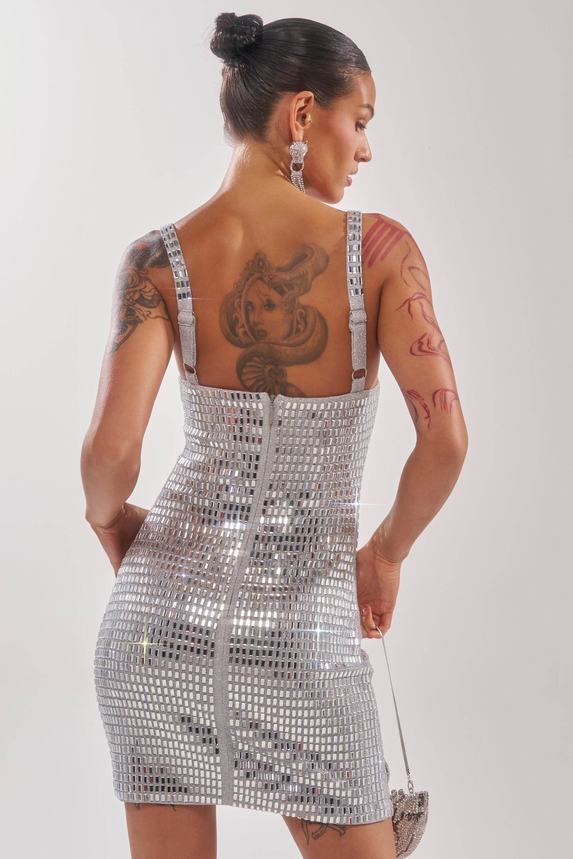 A woman with tattoos wears the BAILEY RHINESTONE MINI DRESS and holds a matching purse. Her hair is in a neat bun as she faces away from the camera, highlighting her back and shoulder tattoos. 5