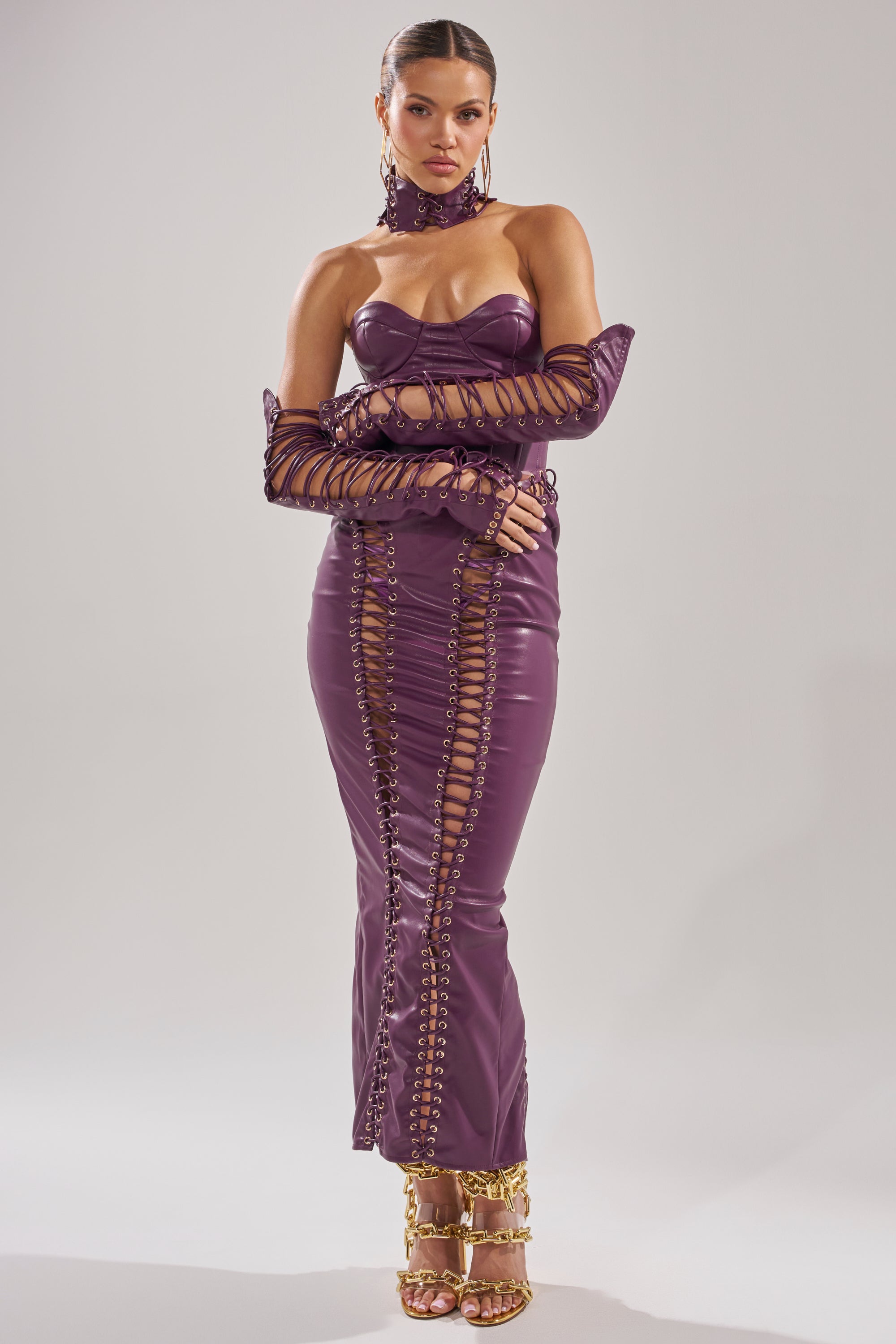 A woman exudes confidence in the WHAT THE HELLY PU MIDI DRESS, a form-fitting purple latex dress with lace-up details, matched with gloves, a choker, statement earrings, and gold high-heeled sandals.