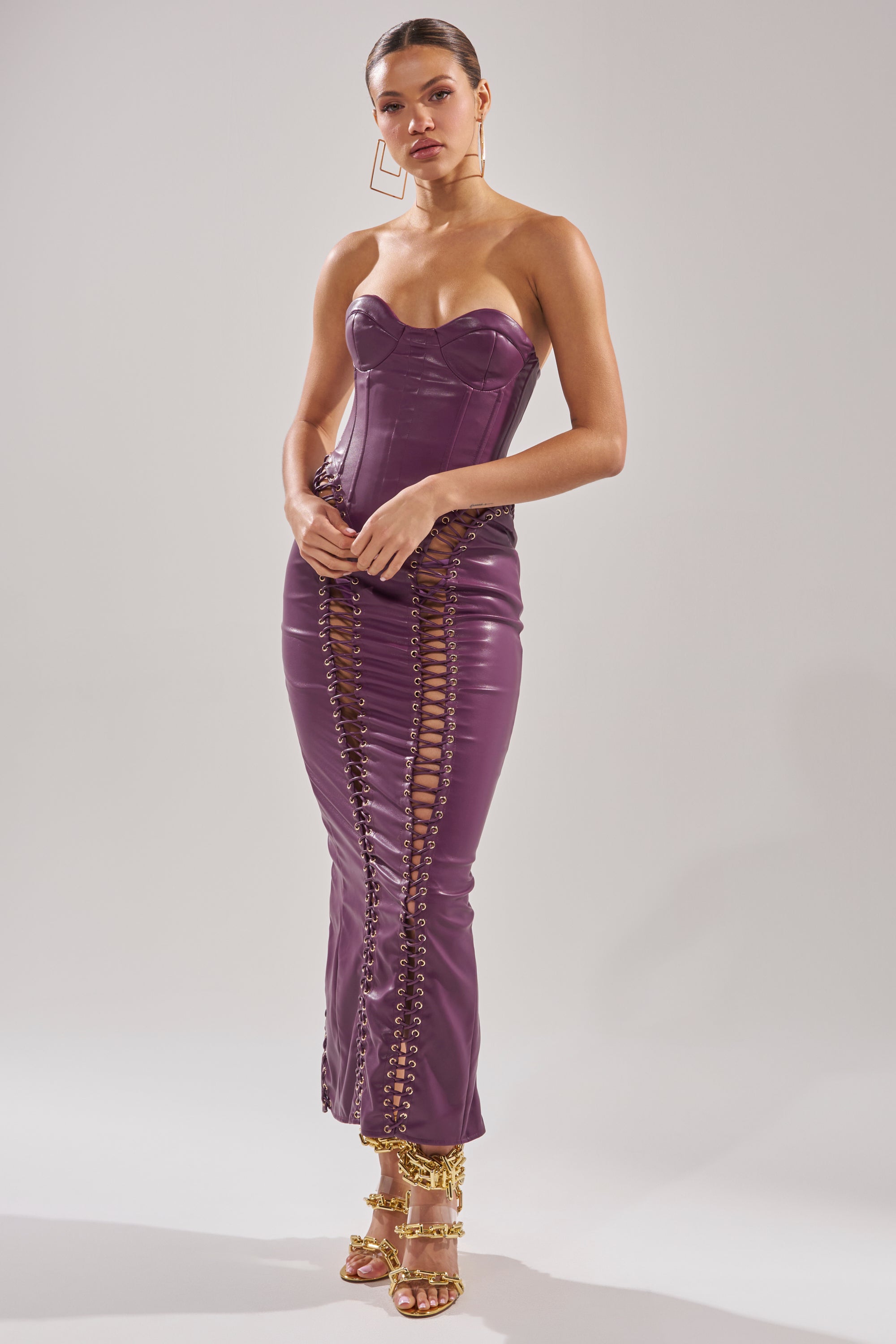 A woman poses against a plain backdrop in the WHAT THE HELLY PU MIDI DRESS—a strapless, form-fitting purple dress with lace-up sides and front—styled with geometric earrings and gold strappy high heels.
