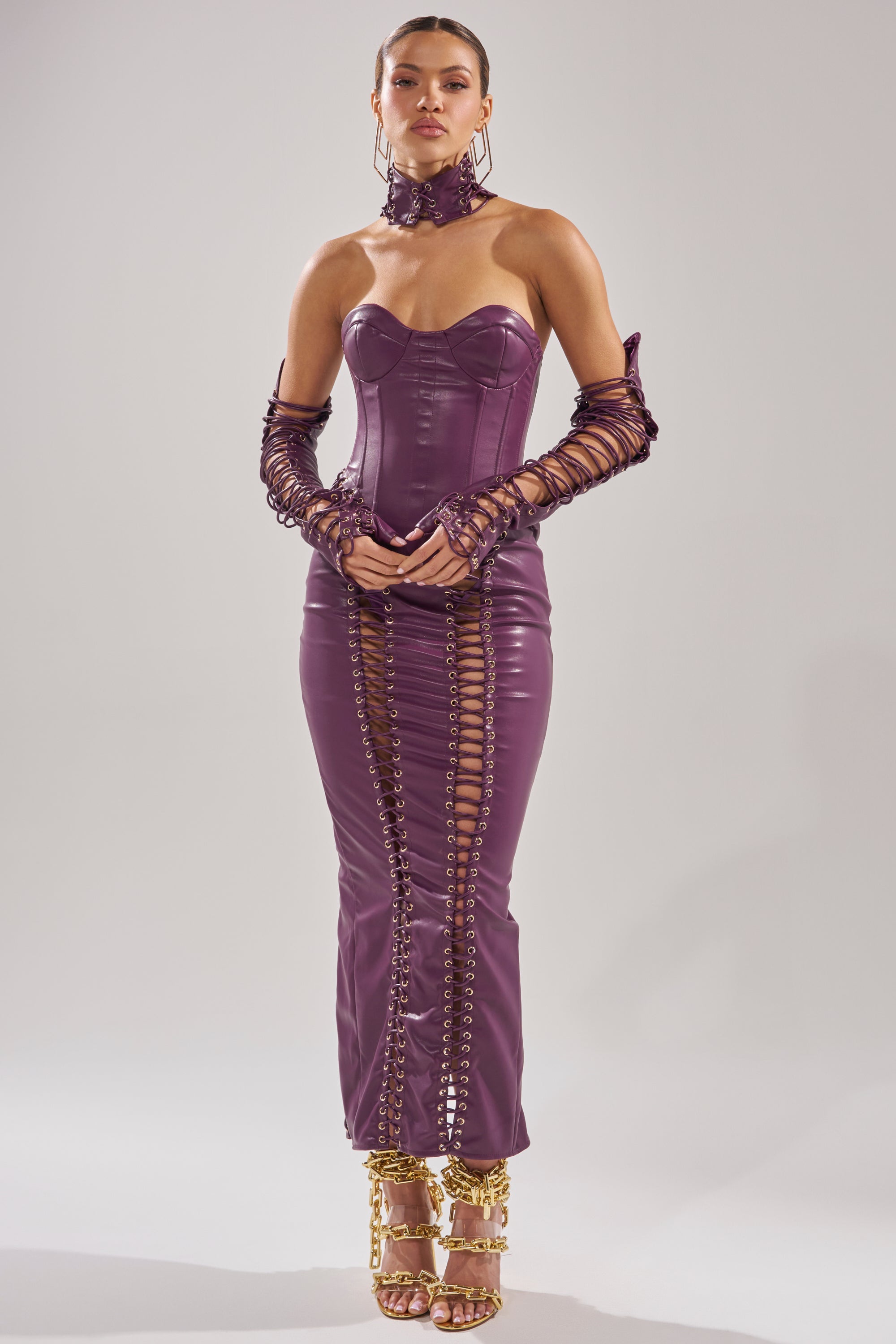 A woman wears the WHAT THE HELLY PU MIDI DRESS, a fitted shiny purple corset dress with lace-up details and matching arm sleeves. She pairs it with a high choker and gold strappy heels, posing against a plain light background.