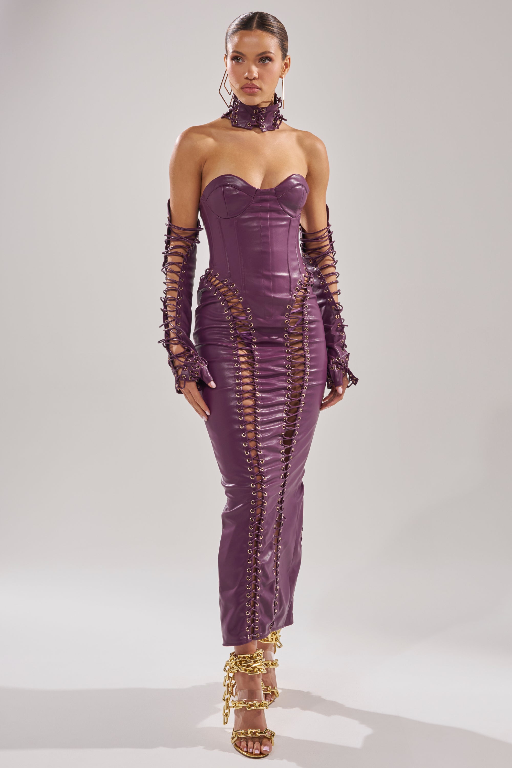 A woman wears the WHAT THE HELLY PU MIDI DRESS, a fitted, strapless purple dress with lace-up details and matching gloves. She pairs it with gold chain heels and a purple choker, standing against a plain light background.