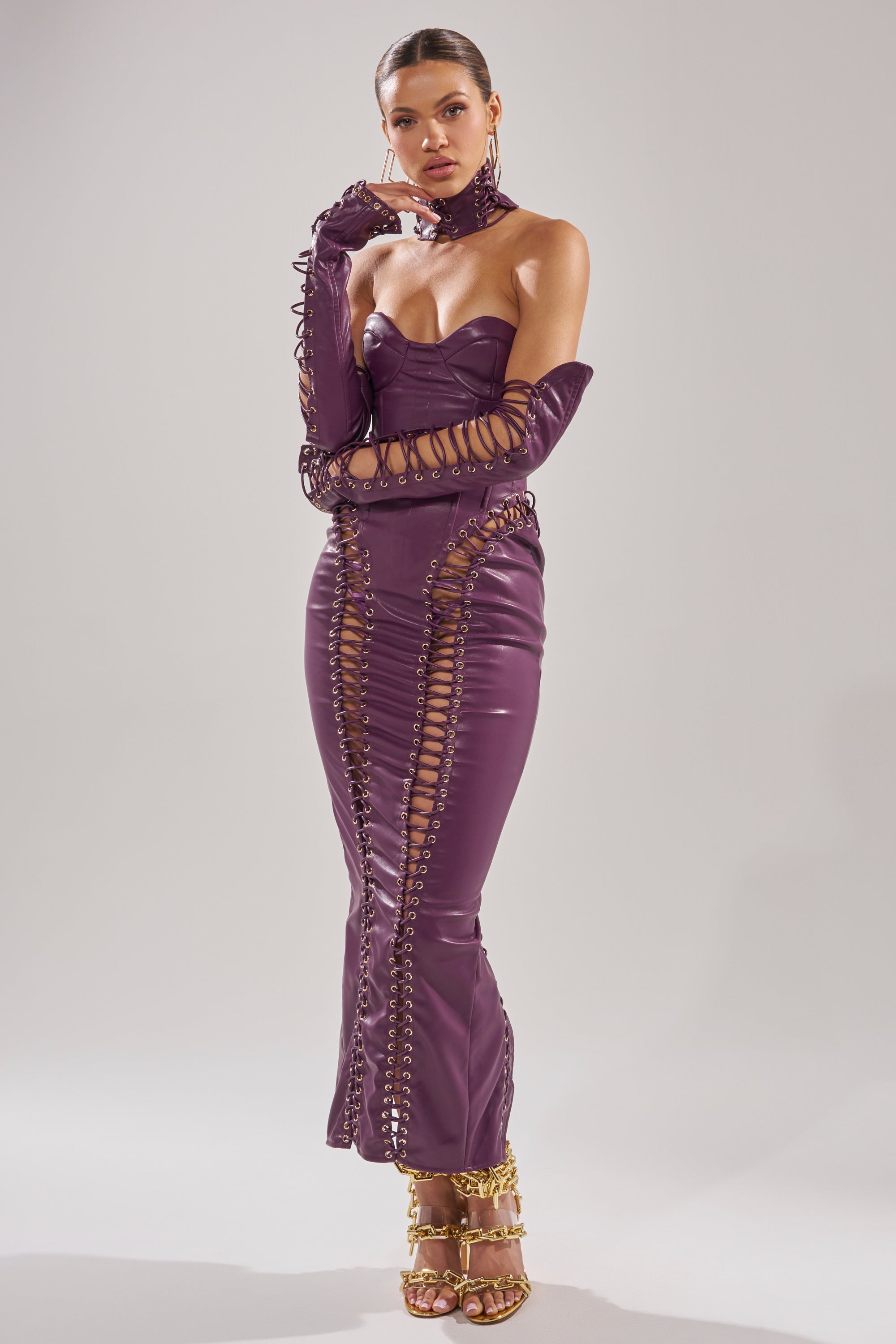 A woman poses in the WHAT THE HELLY PU MIDI DRESS, a fitted deep purple leather style with lace-up details. She accessorizes with matching gloves, a choker, and gold chain-strap high heels against a plain background.