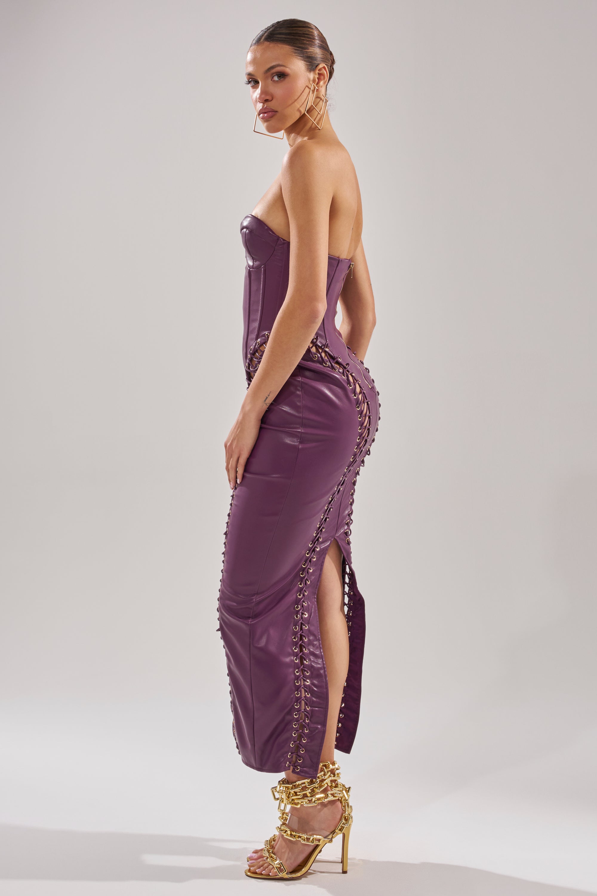 A woman stands sideways in the strapless, form-fitting WHAT THE HELLY PU MIDI DRESS with lace-up sides and a thigh-high slit, paired with gold chain high-heeled sandals and large hoop earrings against a plain light background.