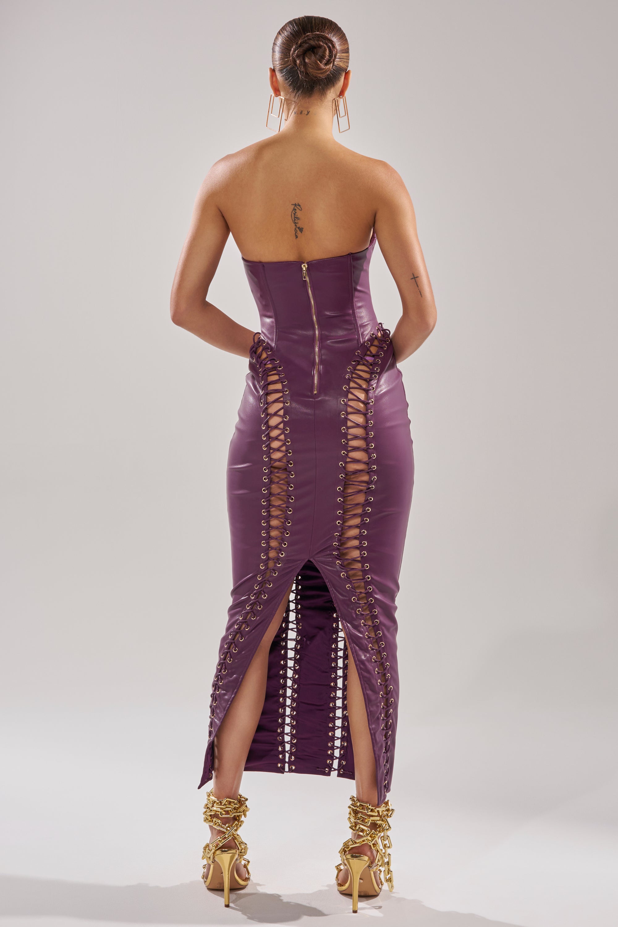 A person stands with their back turned, wearing the WHAT THE HELLY PU MIDI DRESS—strapless, fitted, with lace-up details and slits—paired with gold high heels and a sleek bun hairstyle.