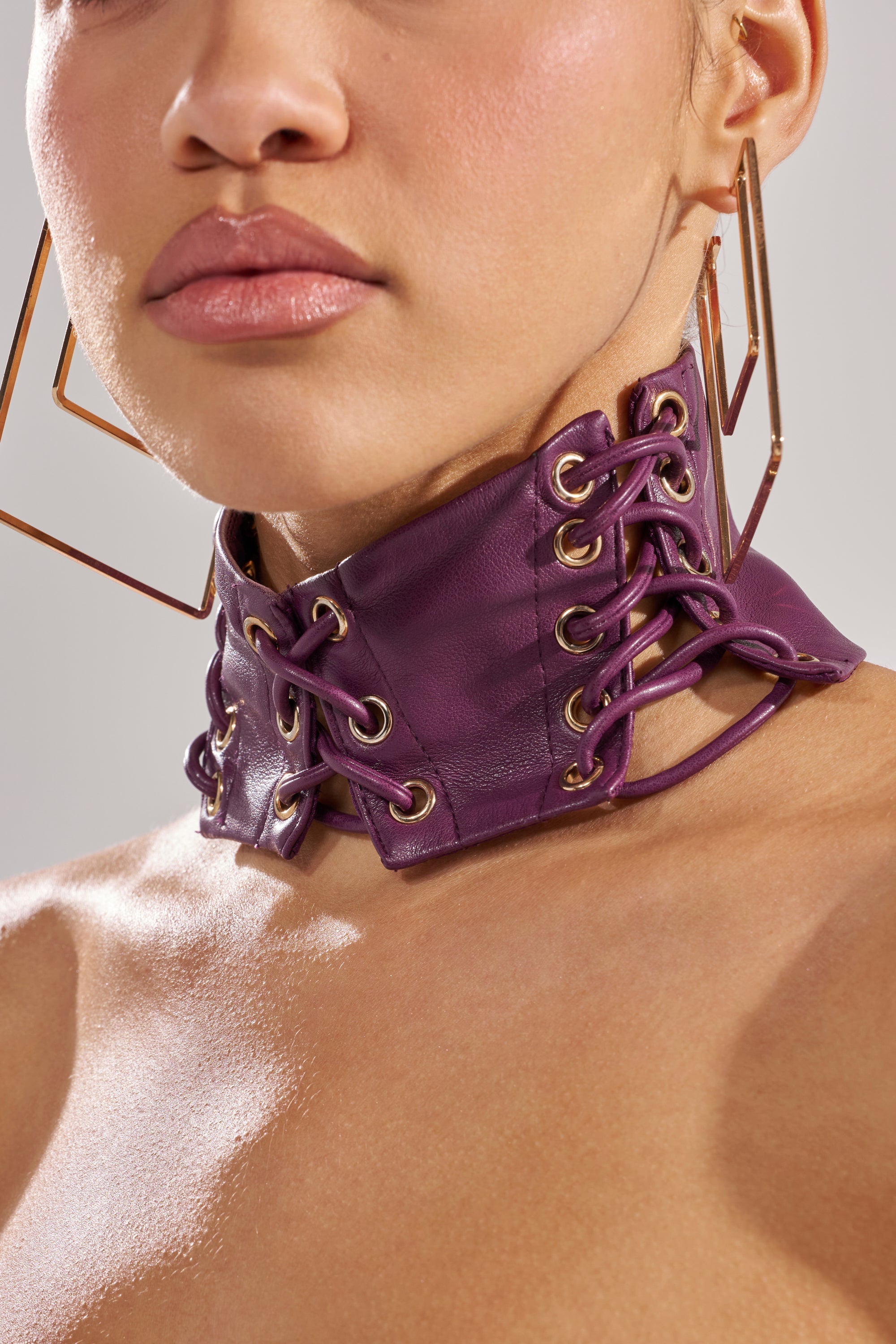 A person wears large geometric earrings and the WHAT THE HELLY CHOKER—a high, wide faux leather choker with gold eyelets, lace-up detailing, covering the neck and part of the upper chest. 0