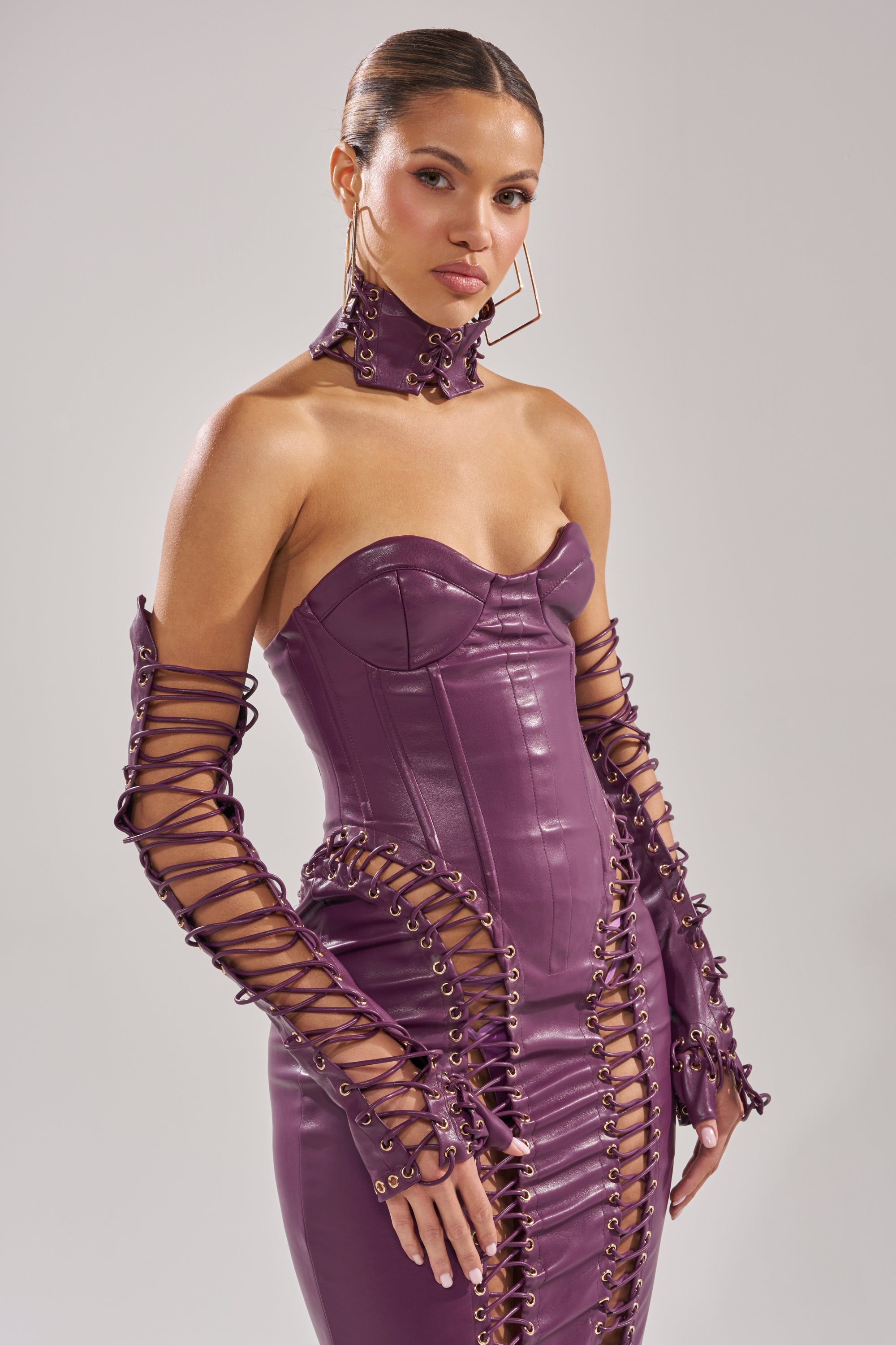 A woman poses in a strapless purple leather dress with lace-up details, matching gloves, and the WHAT THE HELLY CHOKER. Her hair is slicked back, and she wears large geometric earrings against a plain background. 4
