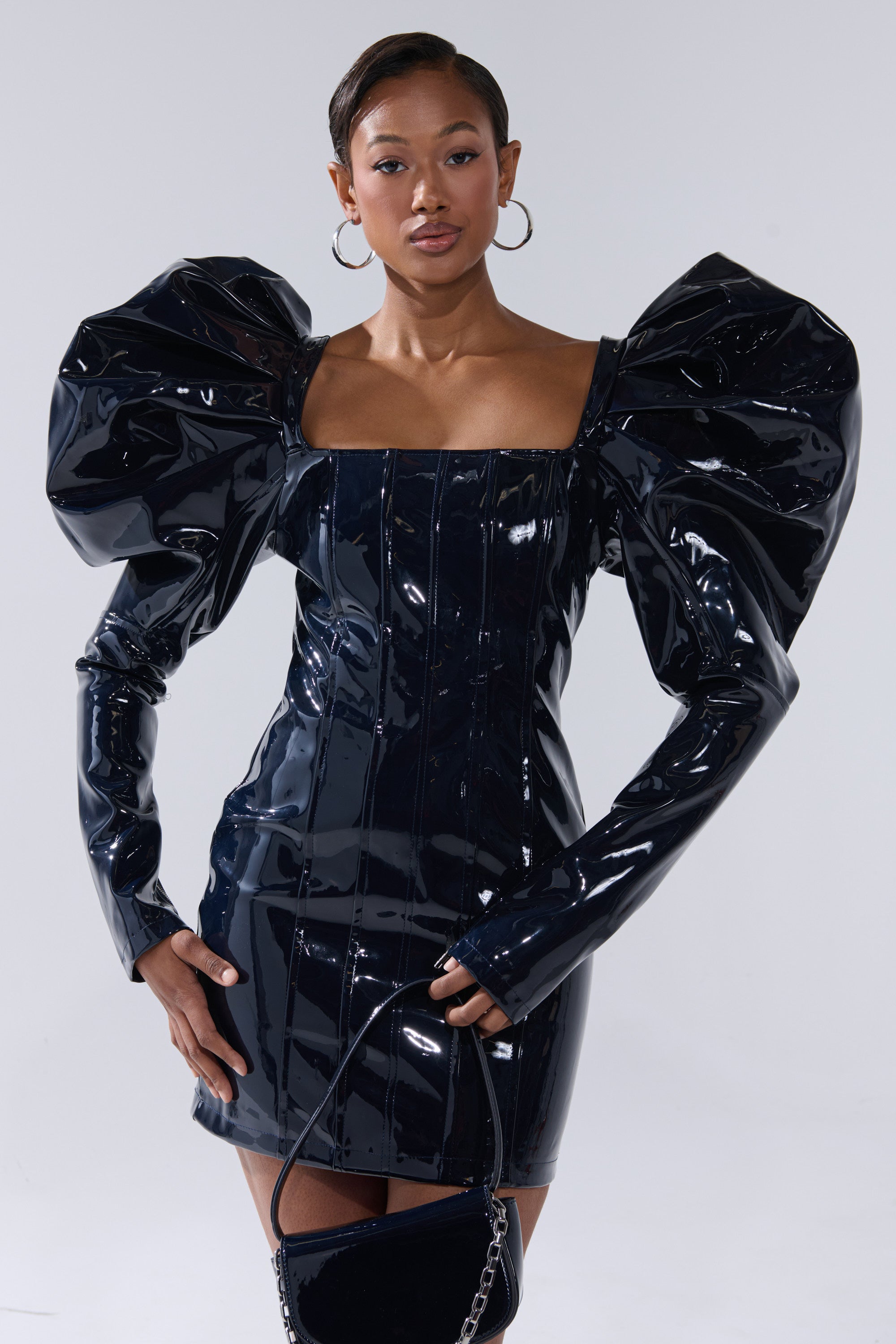 A woman poses confidently in the BADDIES ONLY PATENT MINI DRESS IN NAVY, featuring shiny faux patent leather and dramatic oversized puffed sleeves. She accessorizes with a small black handbag and large hoop earrings against a light background.