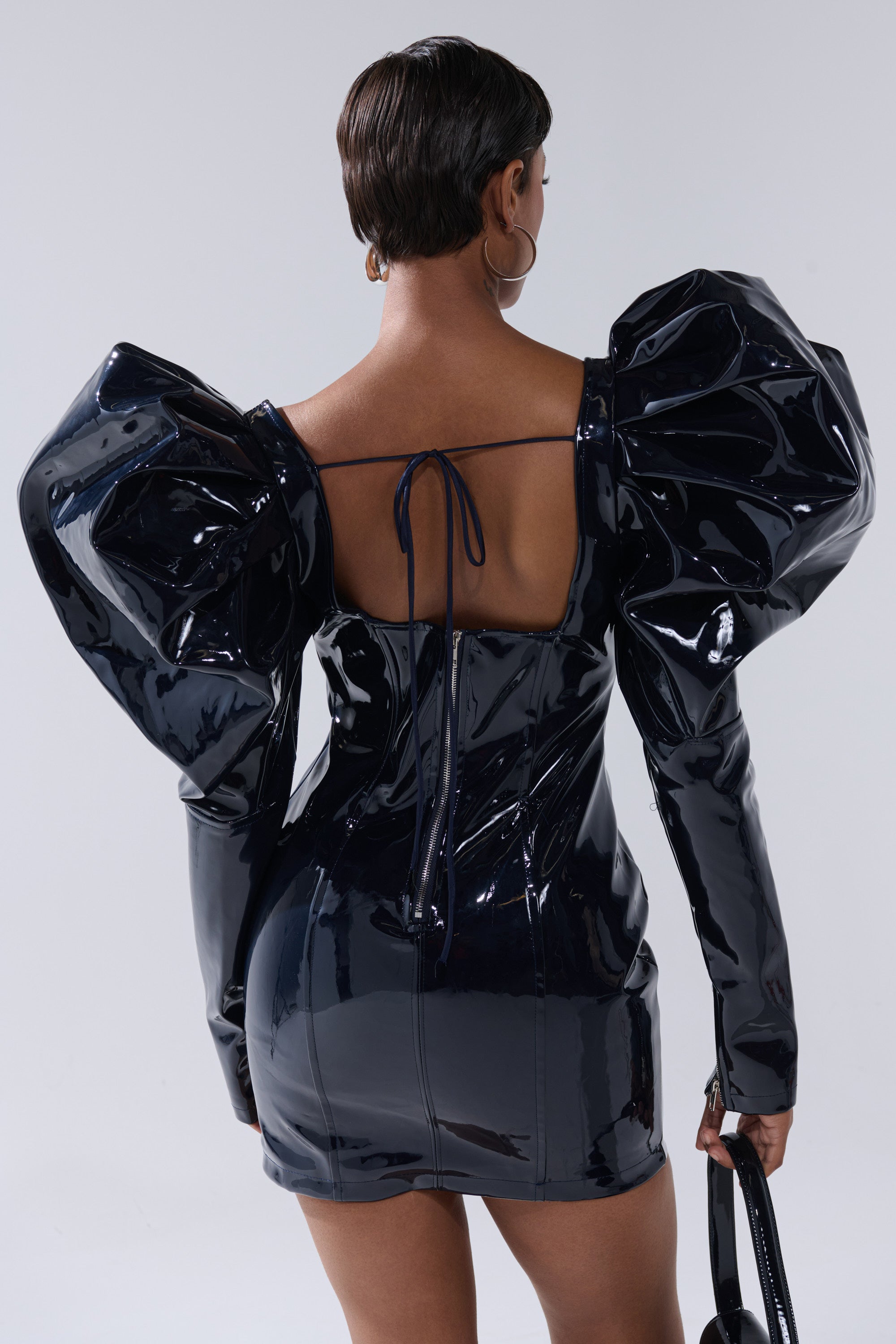 A person with short hair is seen from the back wearing the BADDIES ONLY PATENT MINI DRESS IN NAVY, featuring shiny faux patent leather, dramatic puff sleeves, a tie at the upper back, and holding a small matching bag with hoop earrings.