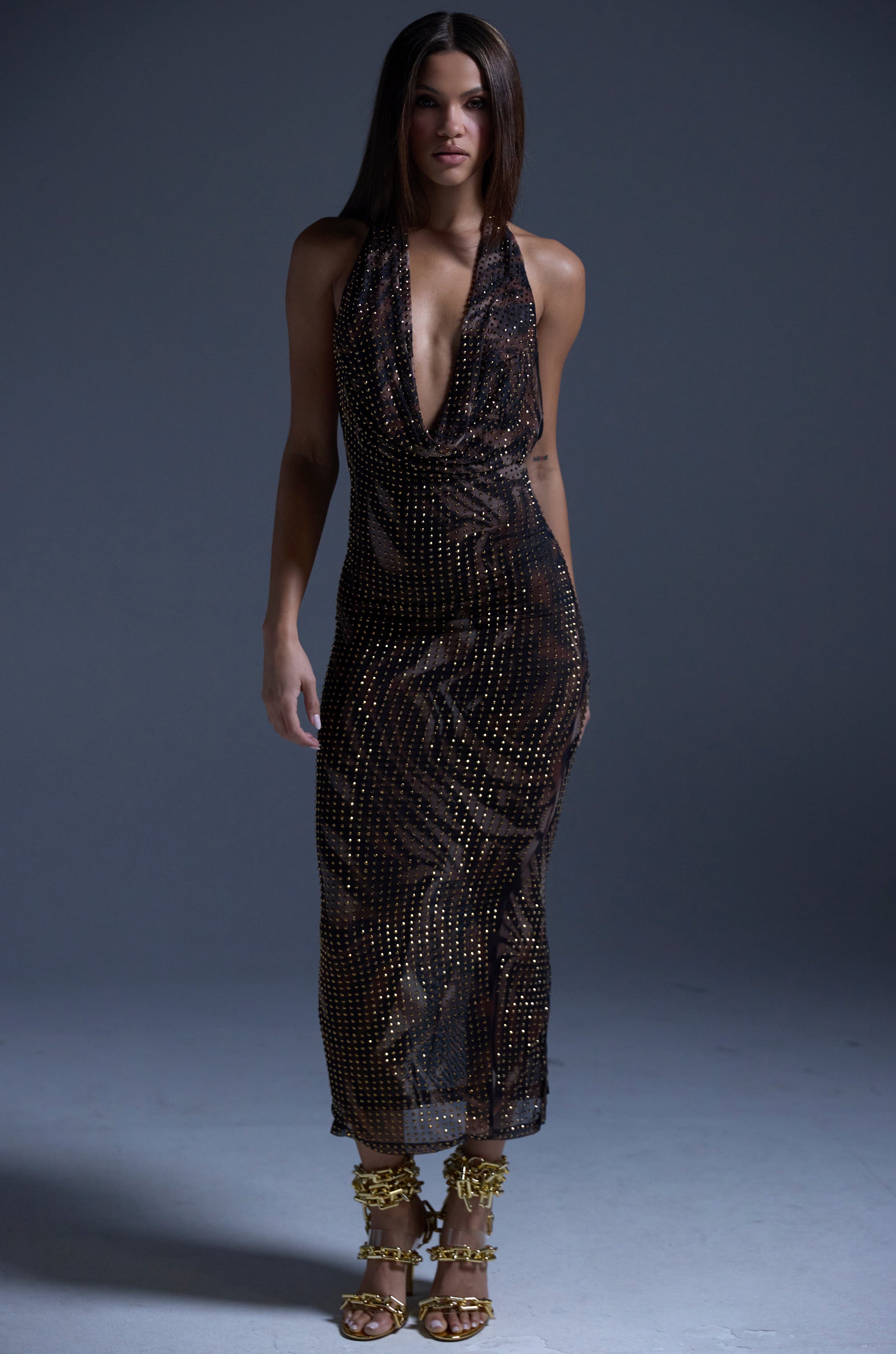 A woman poses against a gray backdrop in the LENNON PRINTED MESH RHINESTONE MIDI DRESS, a sleeveless dark brown style with metallic studs, paired with gold strappy embellished heels. Her straight hair is worn down.