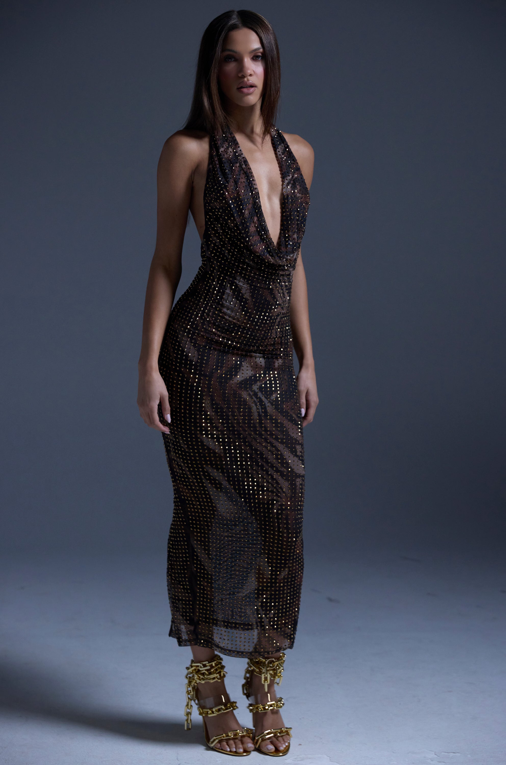 A woman poses against a plain background in the LENNON PRINTED MESH RHINESTONE MIDI DRESS, a sleeveless sheer dark dress with metallic dots, a deep cowl neckline, and gold strappy high heels. Her straight hair is worn loose.