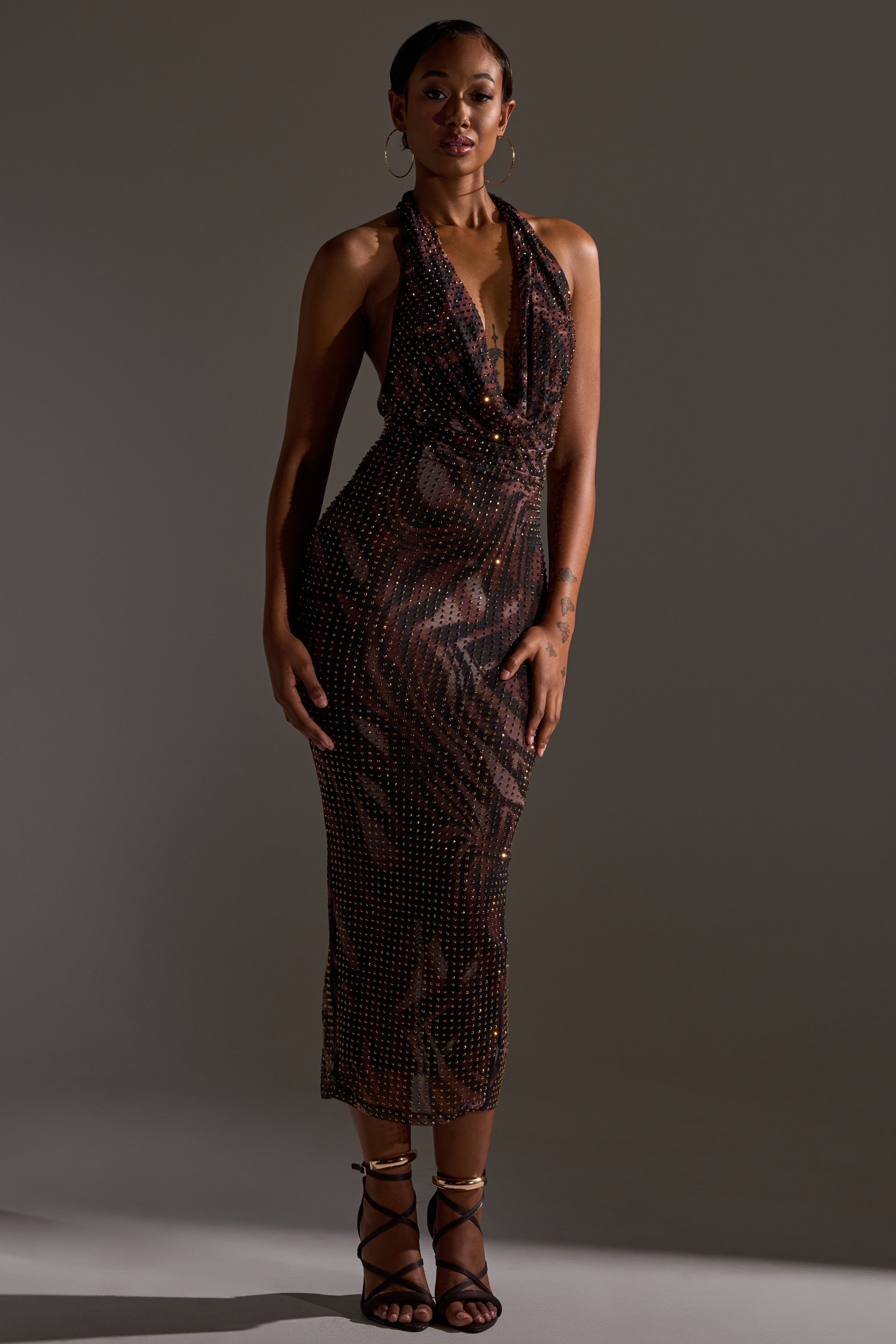 A woman stands confidently against a dark background wearing the LENNON PRINTED MESH RHINESTONE MIDI DRESS, paired with hoop earrings and strappy black heels, her hand resting by her side.