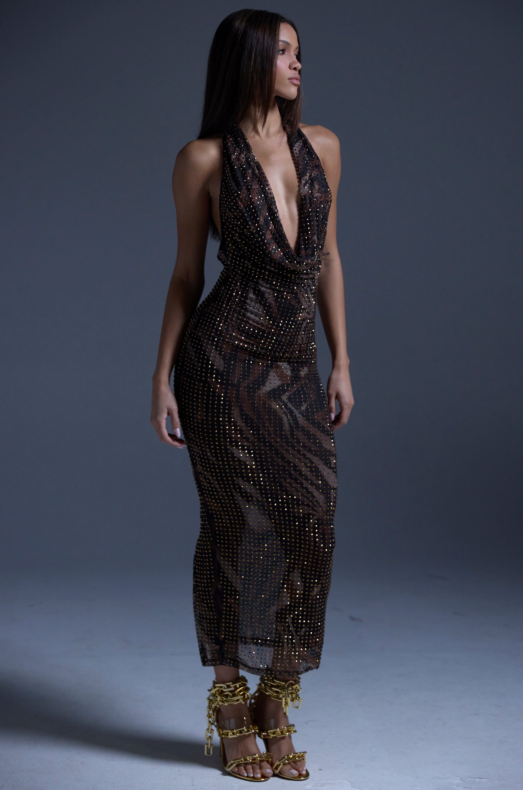 A woman poses against a gray backdrop wearing the LENNON PRINTED MESH RHINESTONE MIDI DRESS, a sleeveless, deep V-neck, sheer brown dress with metallic studs, paired with gold high-heeled sandals featuring ankle chains.