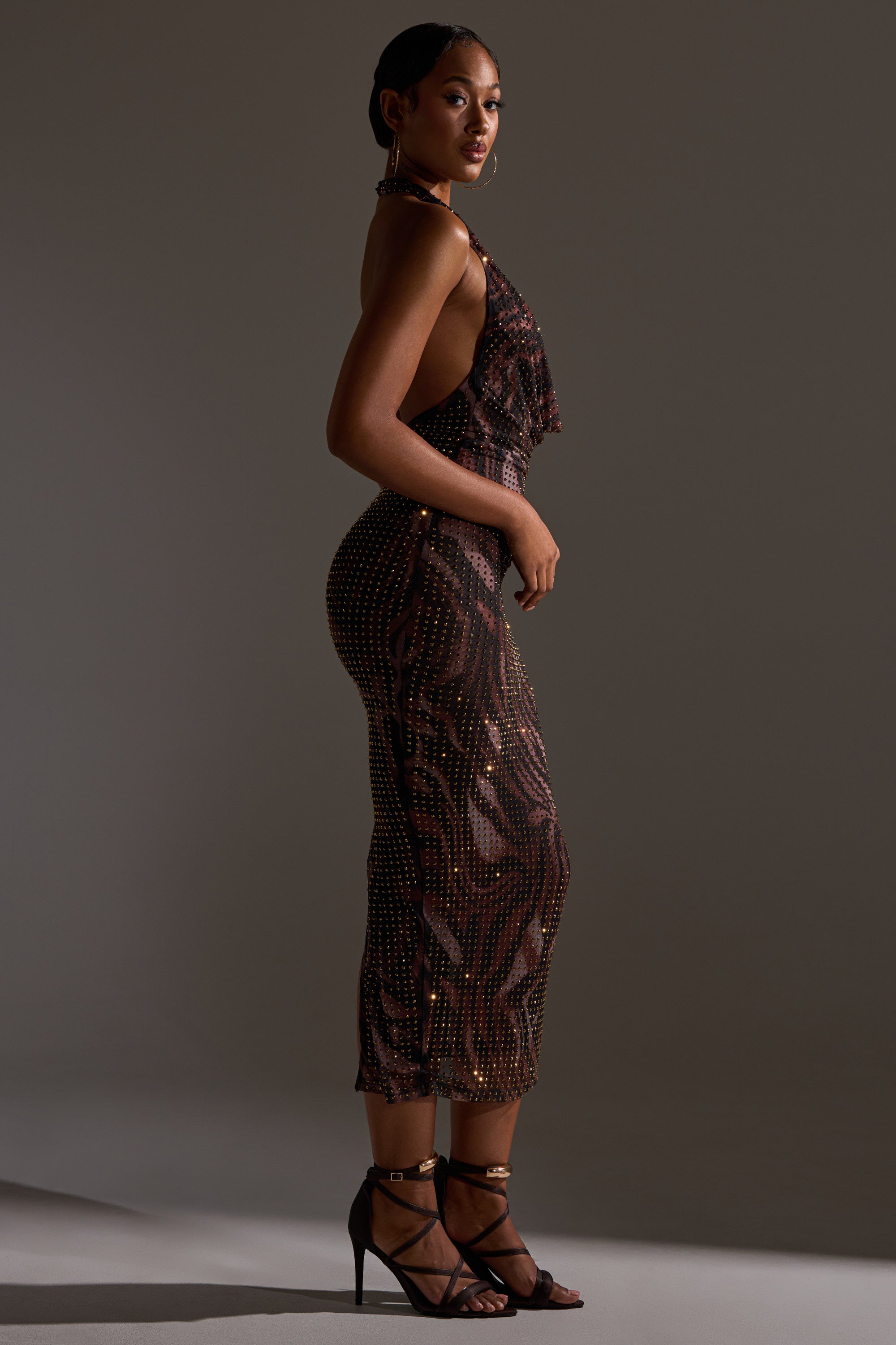 A woman poses in profile against a neutral background, wearing the LENNON PRINTED MESH RHINESTONE MIDI DRESS with animal print and sequins, paired with black strappy high-heeled sandals for a chic, bodycon look.