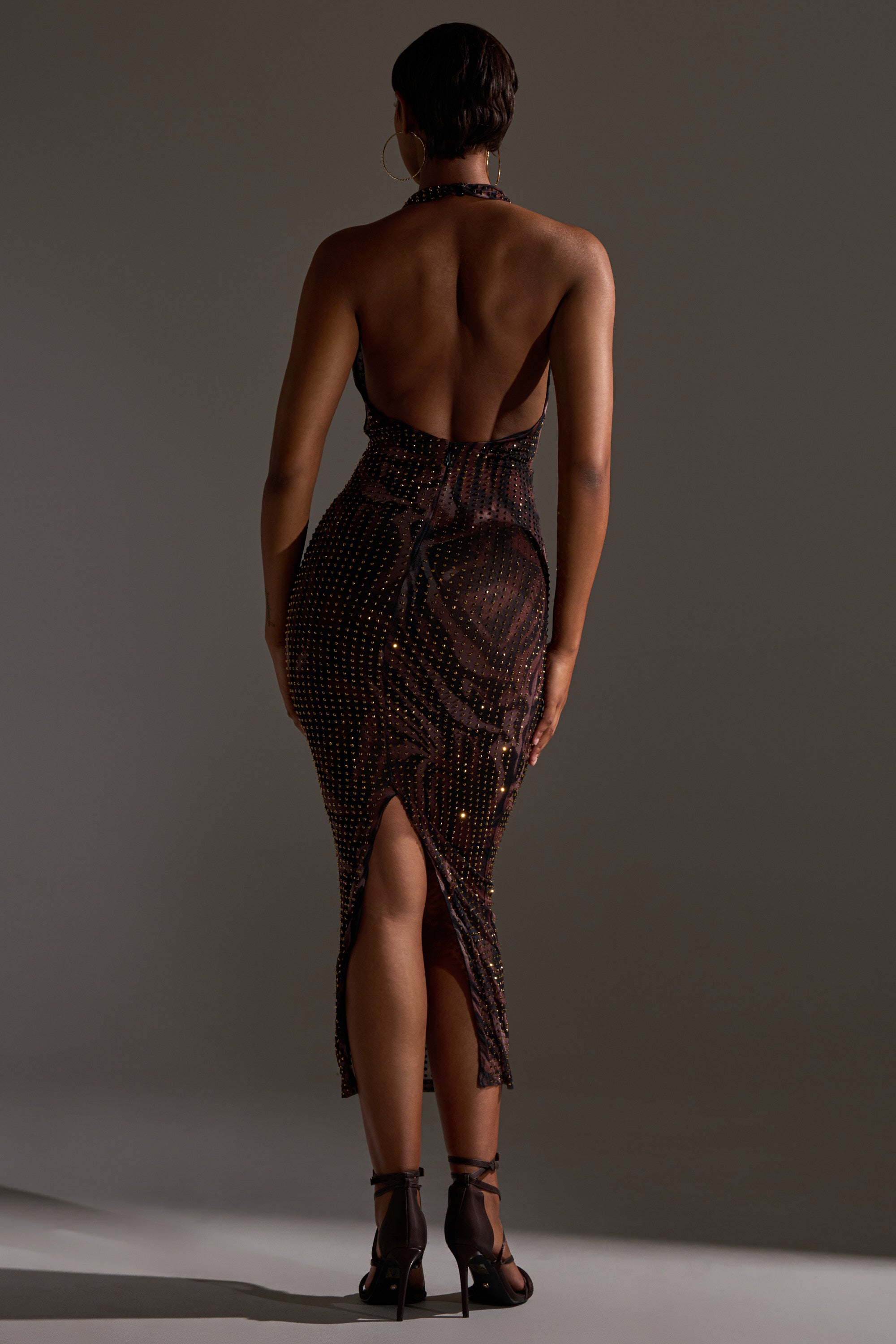 A person faces away, wearing the LENNON PRINTED MESH RHINESTONE MIDI DRESS with an open back, halter neck, and rhinestone details. Their short hair and strappy heels stand out against a softly lit backdrop that highlights the dress’s elegant back slit.