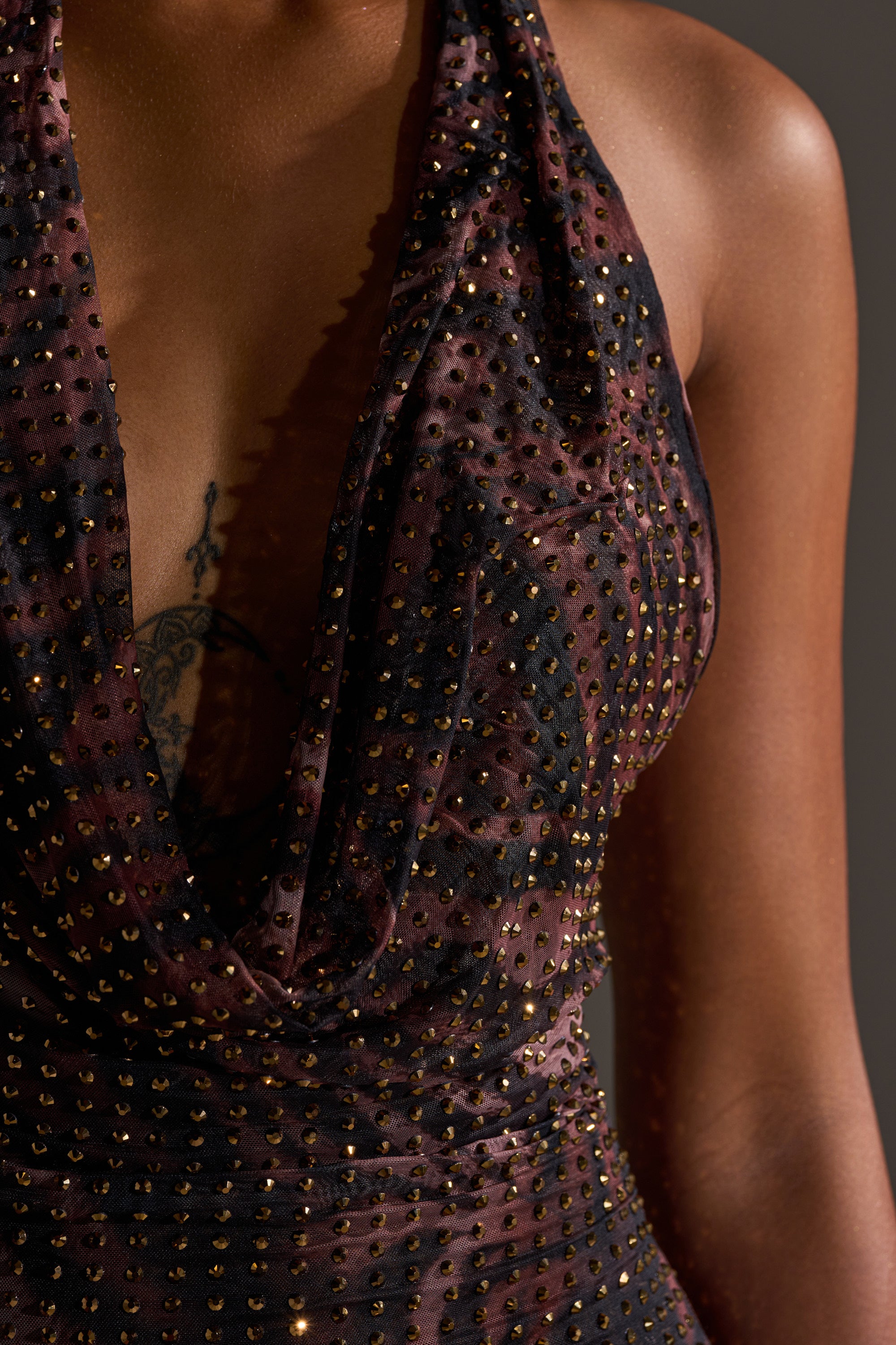 A close-up of a woman wearing the LENNON PRINTED MESH RHINESTONE MIDI DRESS, featuring a sleeveless, sheer cowl neckline and dark fabric embellished with small rhinestones. Her chest tattoo is partially visible against a neutral, blurred background.