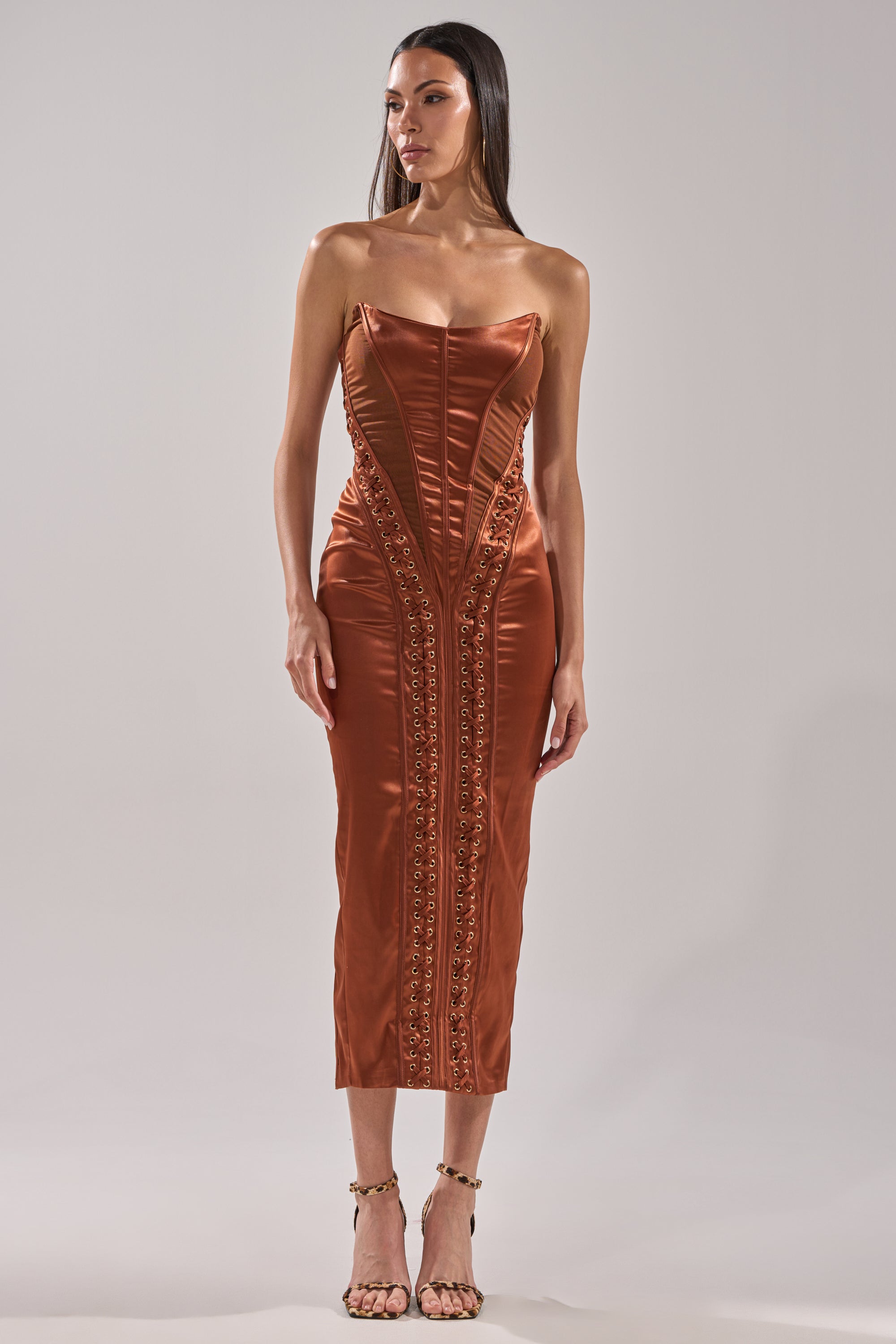 A woman stands against a plain background wearing the WOAH BABY Satin Midi Dress in Chocolate, a form-fitting, strapless dress with structured seams, decorative embellishments, and paired with open-toe heels.