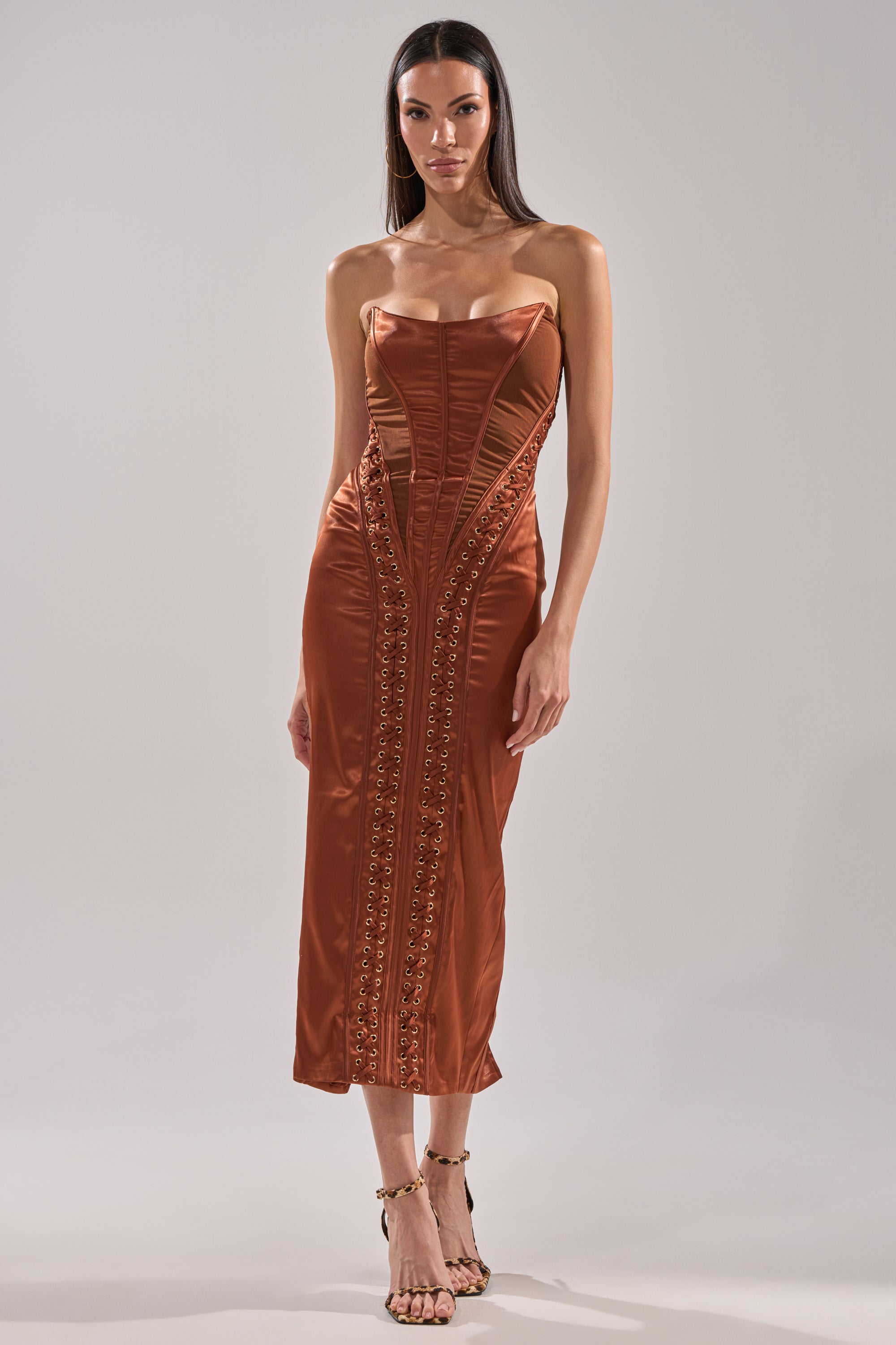 A woman poses against a plain background wearing the WOAH BABY SATIN MIDI DRESS IN CHOCOLATE, a strapless satin dress with decorative vertical stitching and embellishments, paired with strappy high-heeled sandals.