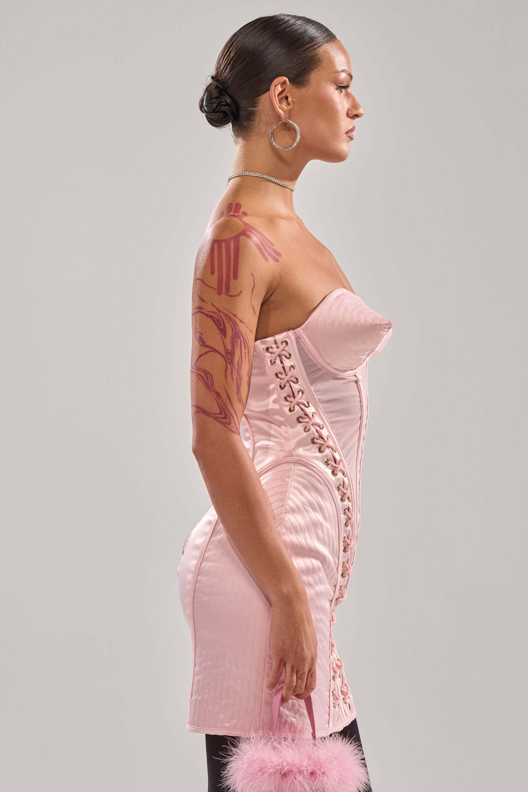 A woman in profile wears the WOAH BABY SATIN MINI DRESS WITH CONE DETAIL—a fitted, light pink mini dress with lace-up sides. She pairs it with hoop earrings, a pearl necklace, slicked-back hair, arm tattoos, and a fluffy pink purse. 7