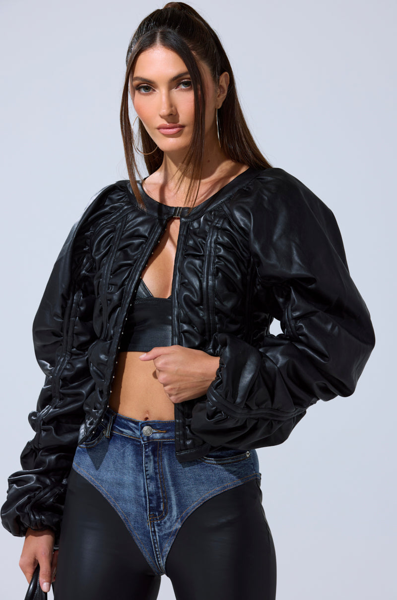 A woman with long brown hair models the AKIRA Label FAUX LEATHER CORSETED MOTO JACKET, pairing it with a black crop top and high-waisted blue denim pants, as she poses confidently against a plain light background.
