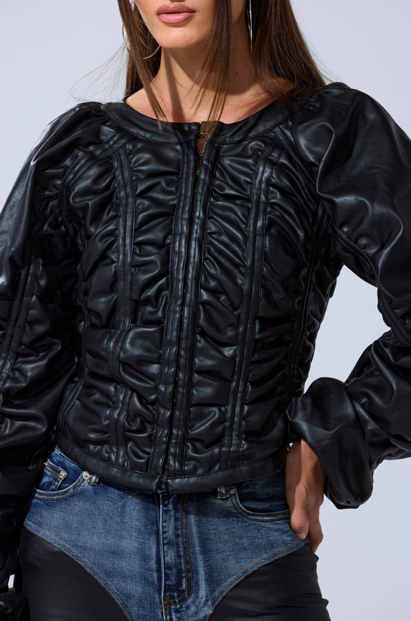 A woman models the AKIRA Label FAUX LEATHER CORSETED MOTO JACKET with voluminous sleeves, styled with blue jeans. She poses against a plain background, one hand on her hip and her face partly visible.