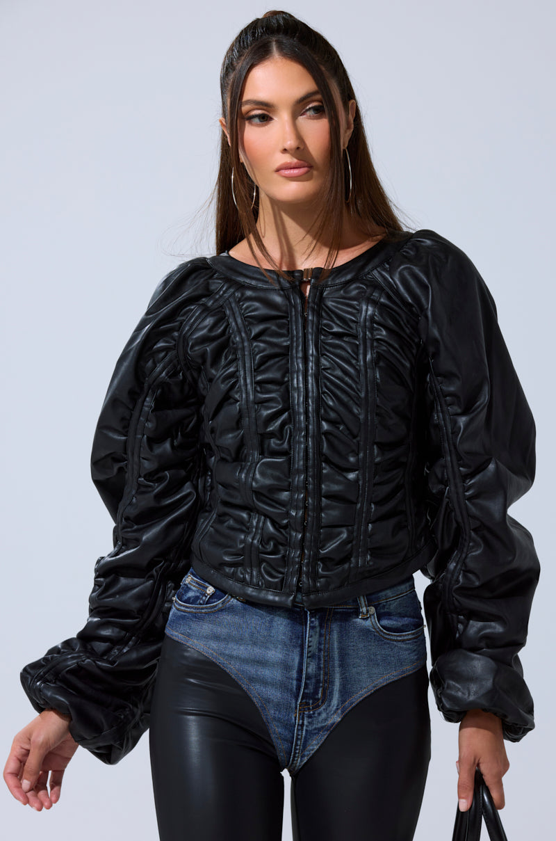 A woman with long brown hair poses confidently against a plain light background, wearing the FAUX LEATHER CORSETED MOTO JACKET with denim high-waisted shorts over black leggings and large hoop earrings.