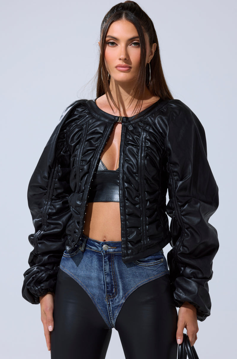 Wearing the FAUX LEATHER CORSETED MOTO JACKET over a black bralette and distinctive high-waisted pants, she styles her long brown hair with hoop earrings and carries a handbag.