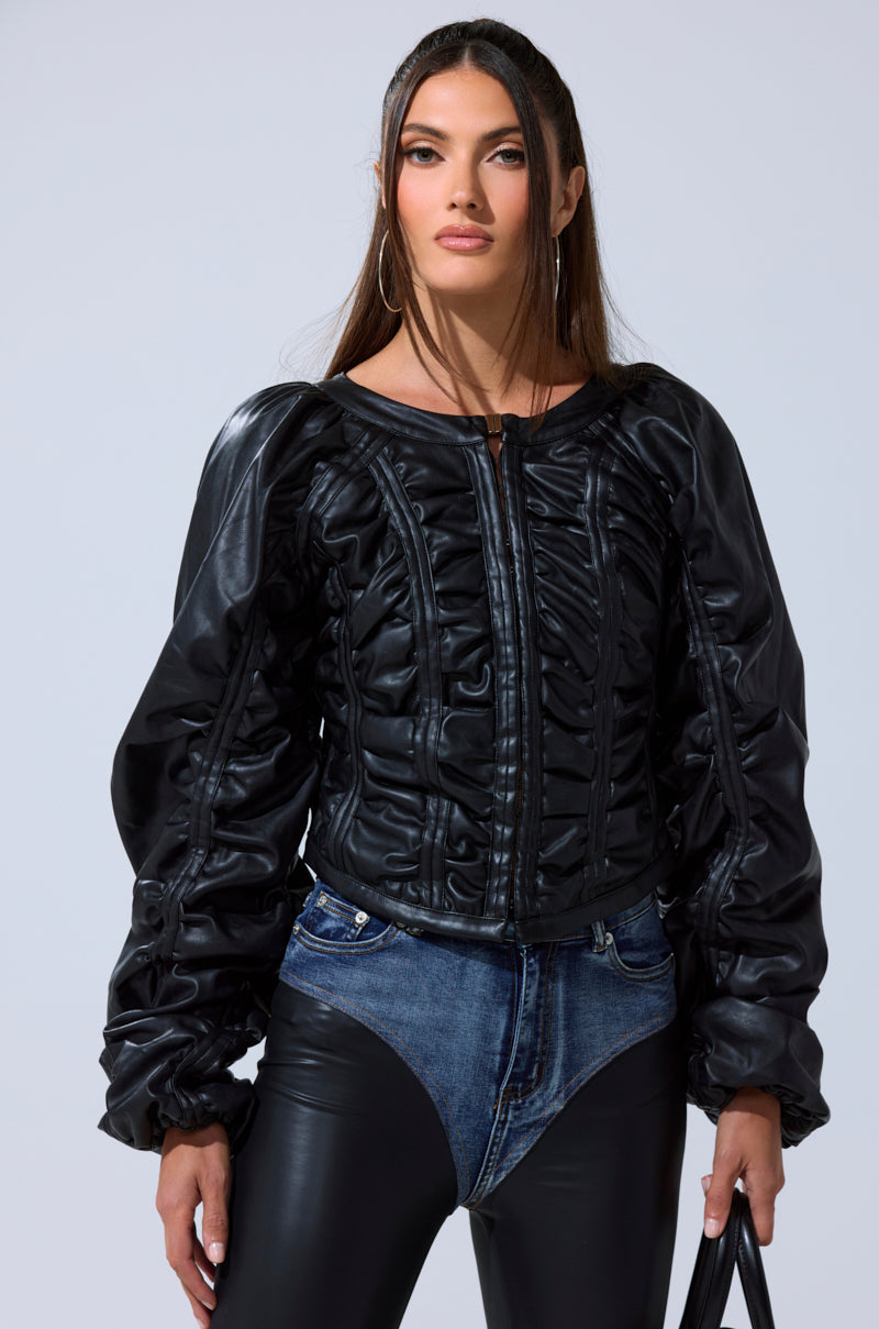 A woman wears the FAUX LEATHER CORSETED MOTO JACKET with voluminous sleeves, styled with black leather pants featuring a denim waistband. She has long, straight hair and carries a black bag.
