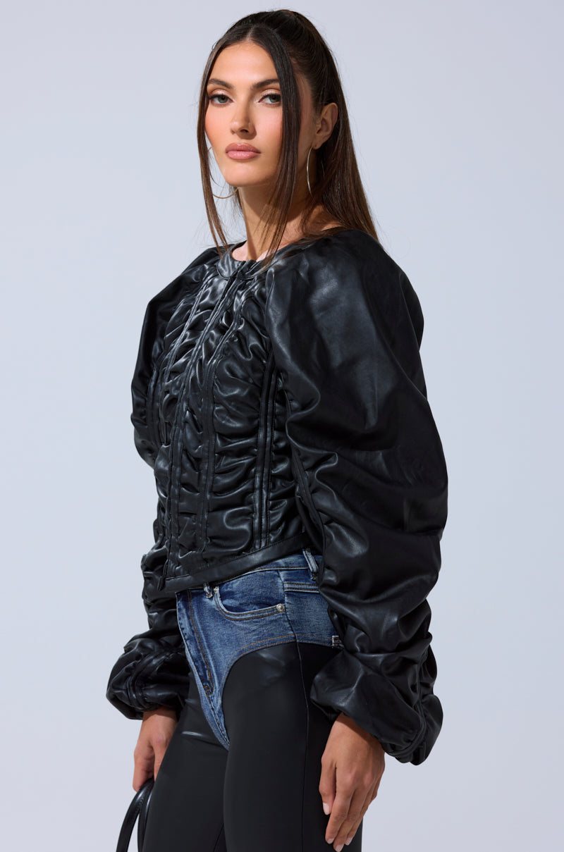 A woman with long brown hair wears an AKIRA Label FAUX LEATHER CORSETED MOTO JACKET with voluminous sleeves and two-tone jeans. She holds a bag and poses confidently against a plain light background, looking at the camera.