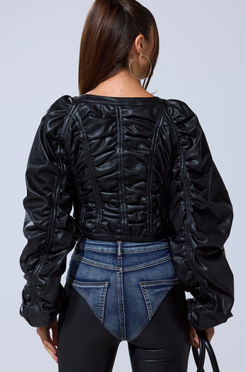 A woman with long brown hair in a ponytail is seen from behind, wearing the FAUX LEATHER CORSETED MOTO JACKET with puffed sleeves and high-waisted jeans that have a unique V-shaped denim design over black leggings.
