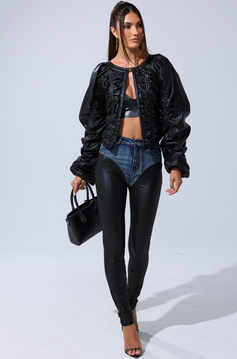 A woman in the AKIRA Label FAUX LEATHER CORSETED MOTO JACKET, a black crop top, high-waisted jeans with black leather leggings, black heeled sandals, and a black handbag stands against a plain light gray background.