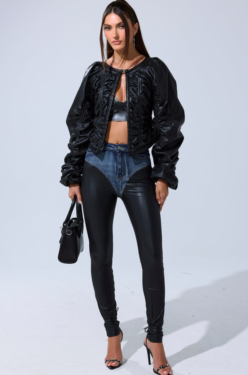 In a studio, a woman wears a black crop top, the FAUX LEATHER CORSETED MOTO JACKET as outerwear, high-waisted denim and leather leggings, strappy heels, and carries a black handbag.