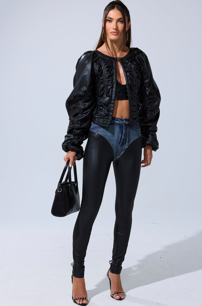 A woman stands against a plain background wearing an AKIRA Label FAUX LEATHER CORSETED MOTO JACKET, black bandeau top, high-waisted black pants with denim briefs detail, black high-heeled sandals, and holding a black handbag.
