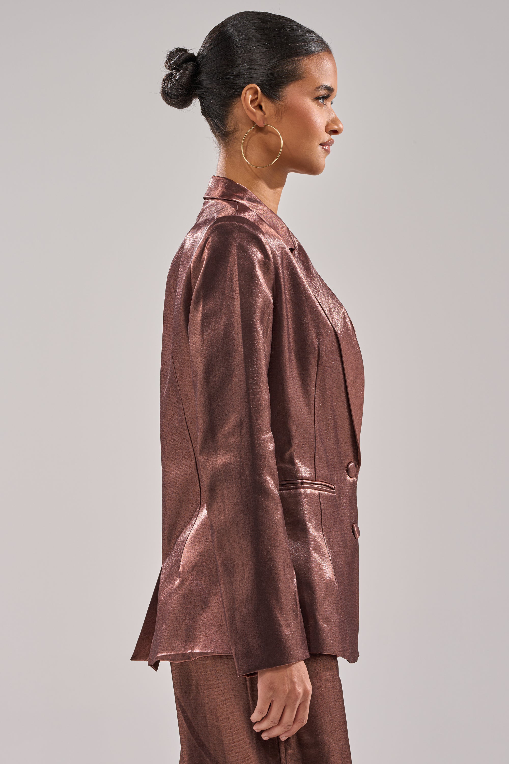 A woman in profile wears the STAY CLOSE IRIDESCENT BLAZER with matching pants, her hair in a neat bun, large hoop earrings, set against a plain light gray background. 2