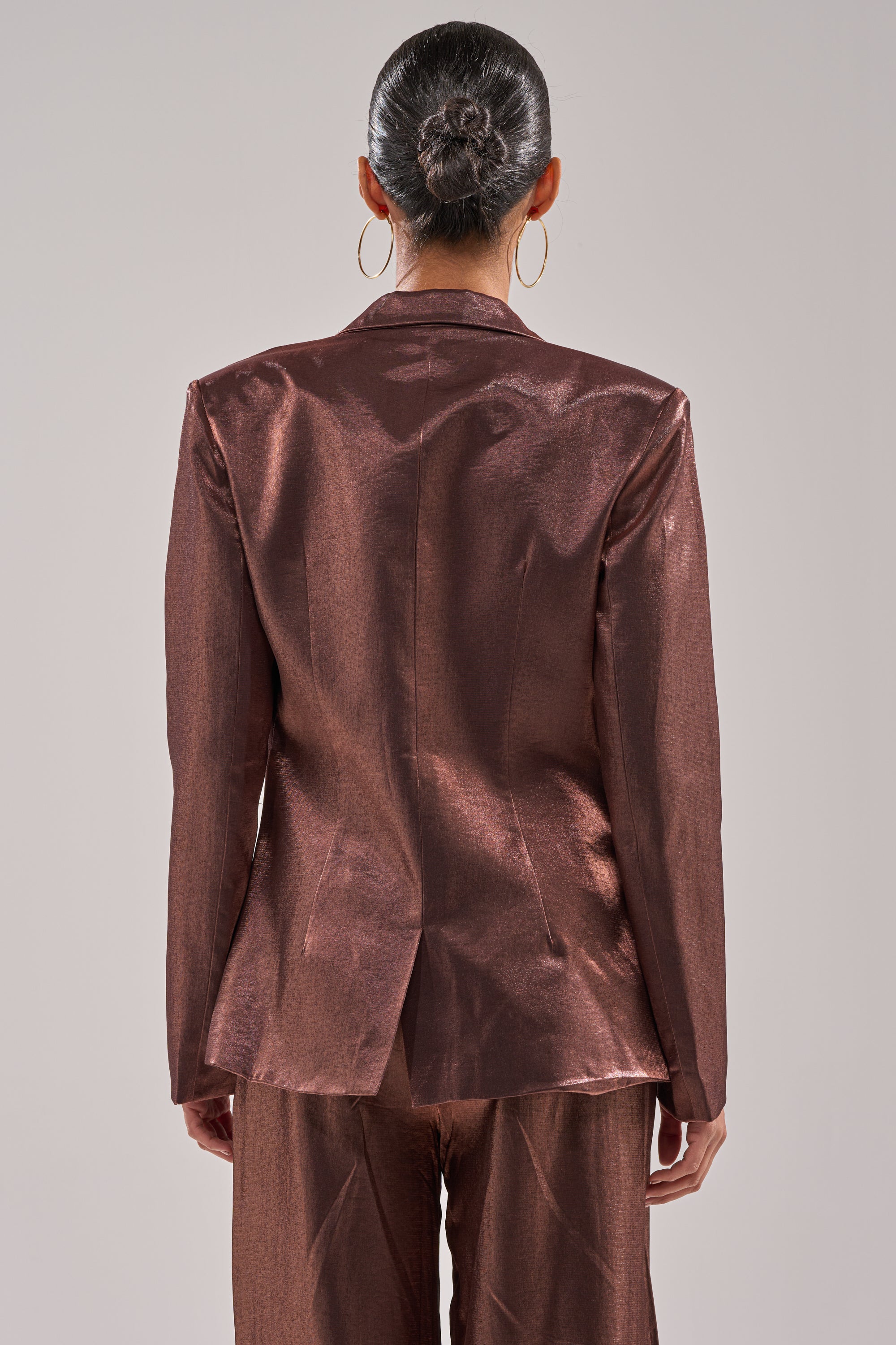 A person with dark hair in a neat bun and large hoop earrings stands facing away, wearing the STAY CLOSE IRIDESCENT BLAZER with matching bronze-brown pants. 4