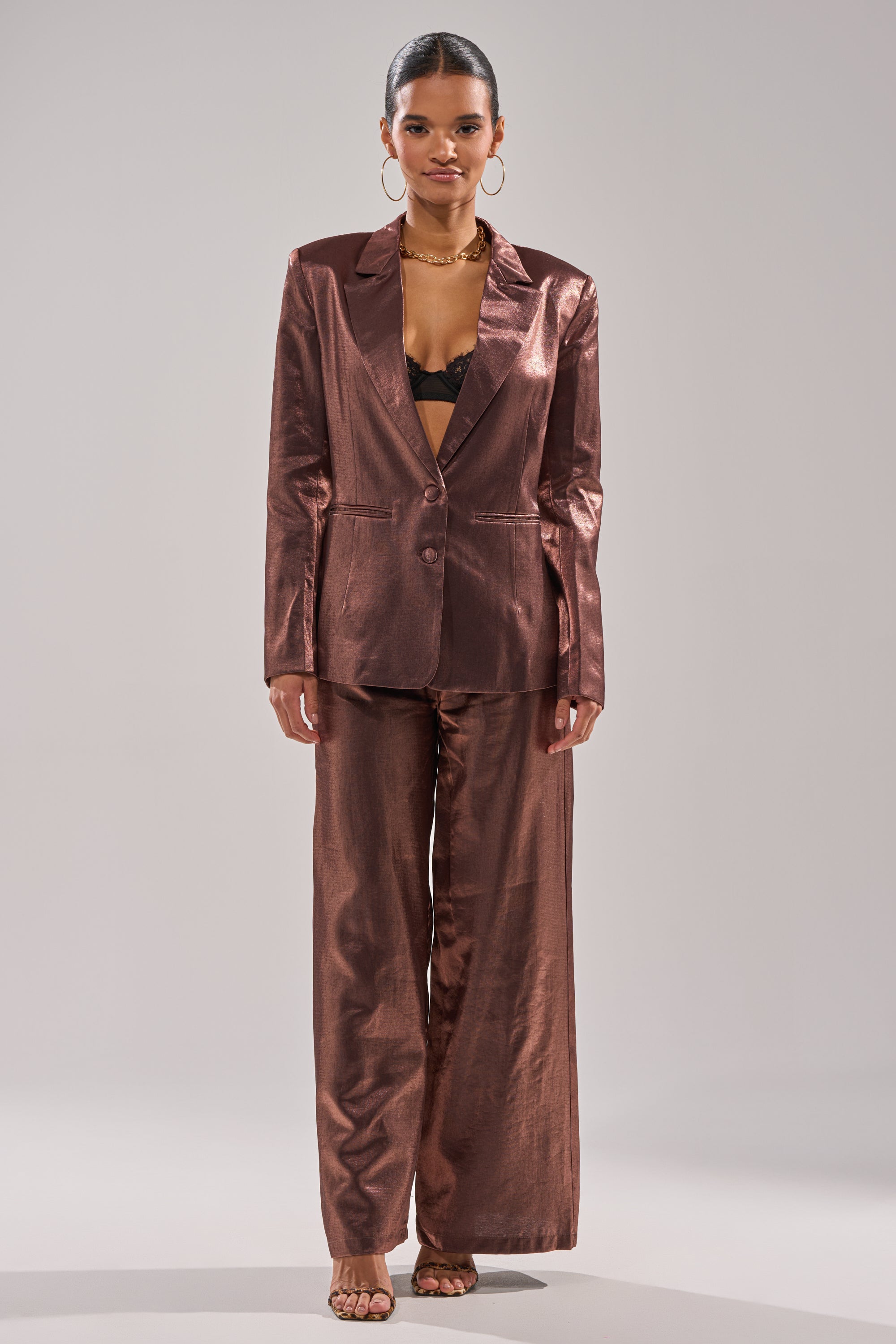 A woman faces forward in the STAY CLOSE IRIDESCENT BLAZER with wide-leg trousers, an open front revealing a black lace bralette, accessorized with hoop earrings, layered necklaces, and heeled sandals. 6