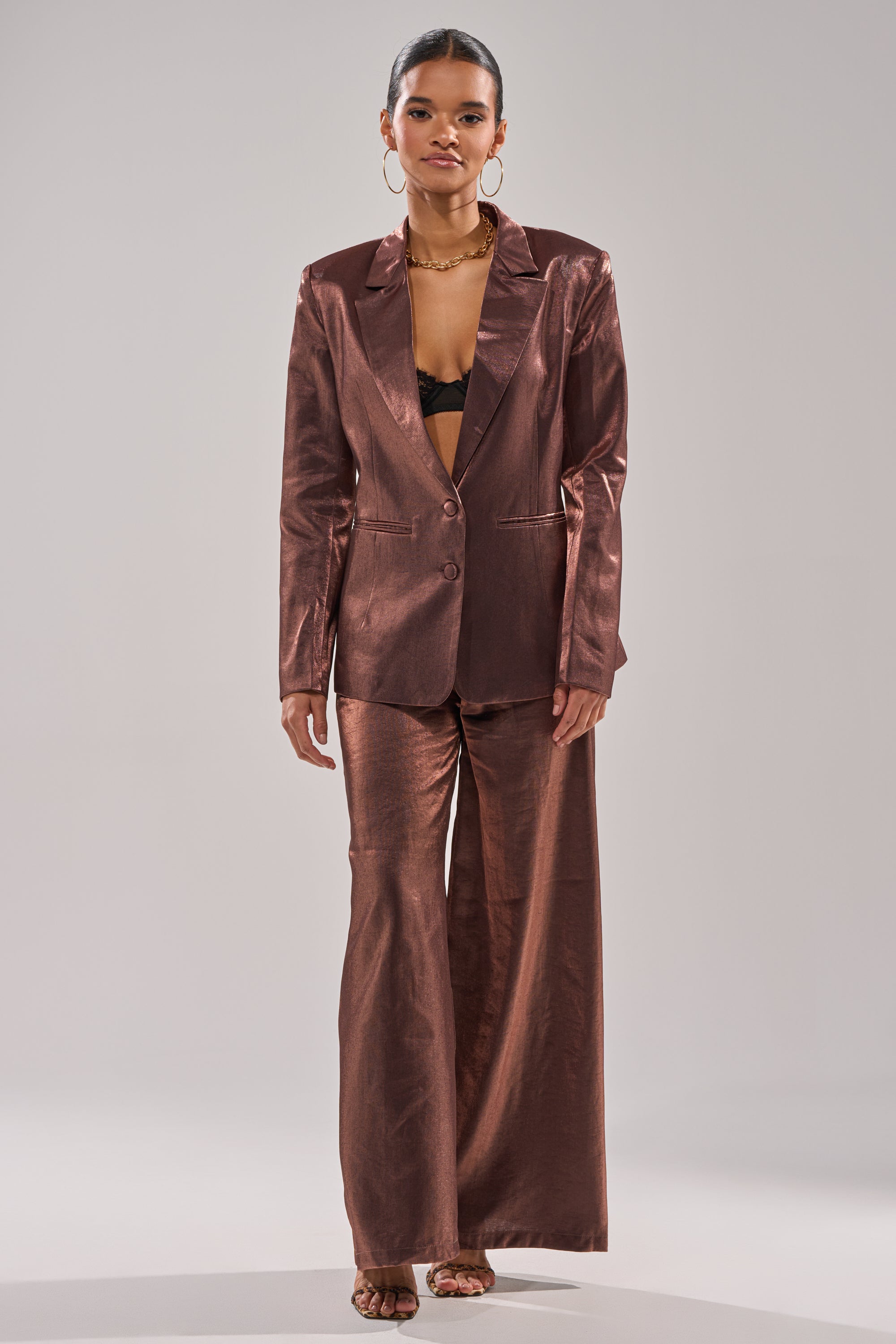 A woman wears the STAY CLOSE IRIDESCENT BLAZER with wide-leg trousers and a black bralette, accessorized with hoop earrings, a necklace, and strappy sandals. 7